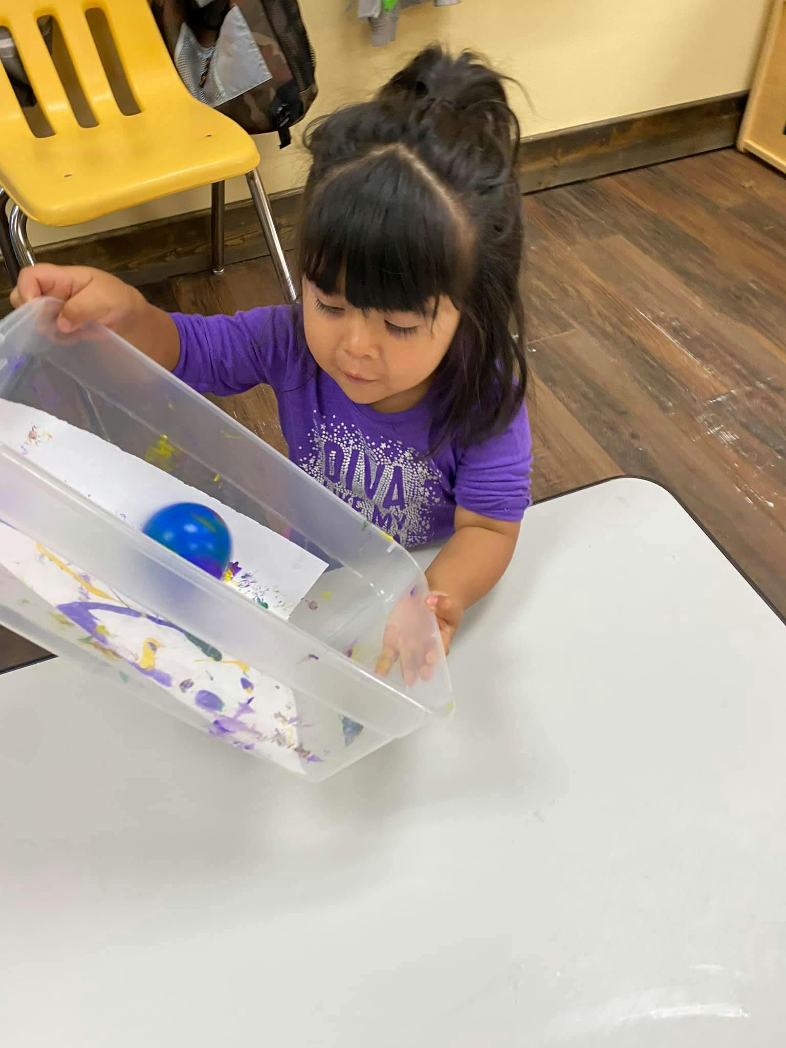 Child holding a clear container with a blue ball and paper with paint on paper