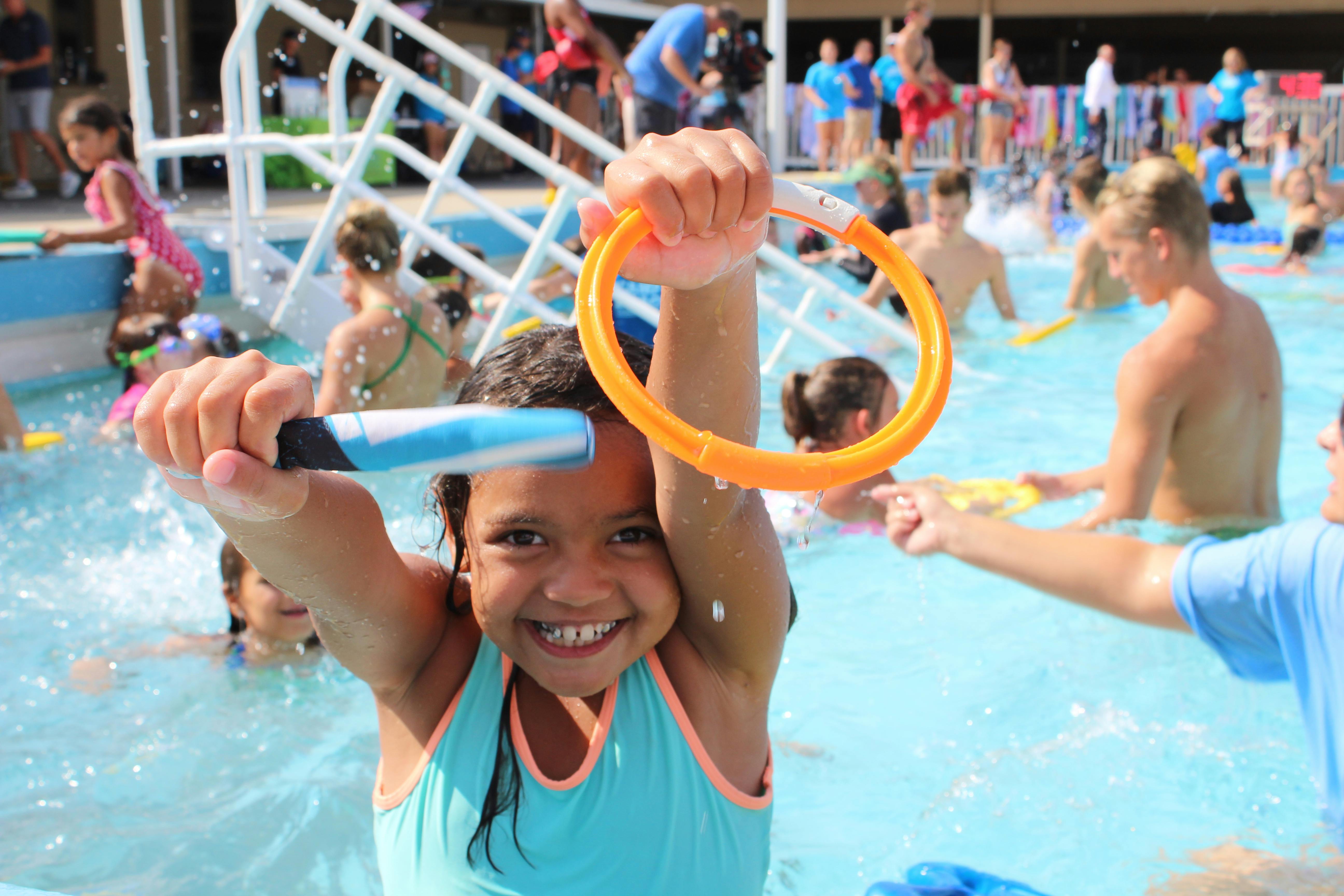 Child in a pool holding up a blue toy and an orange ring