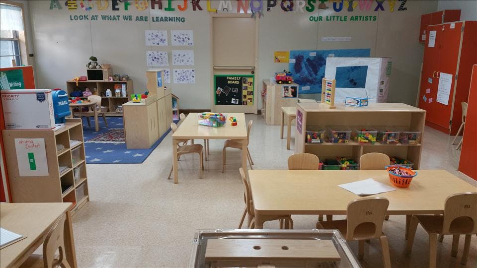 An empty classroom with small tables and chairs, shelves with toys, and wall decorations
