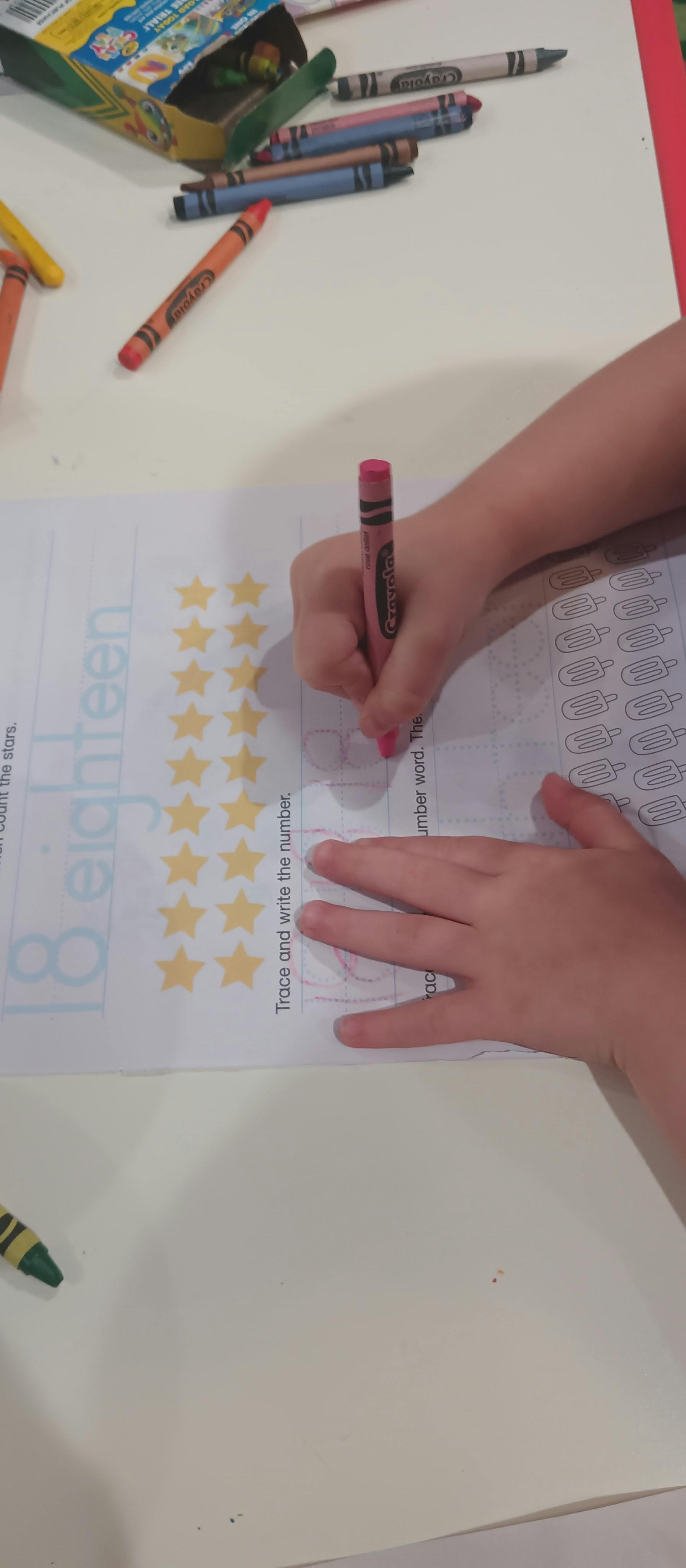 Child's hand holding a pink crayon, writing on a worksheet with stars