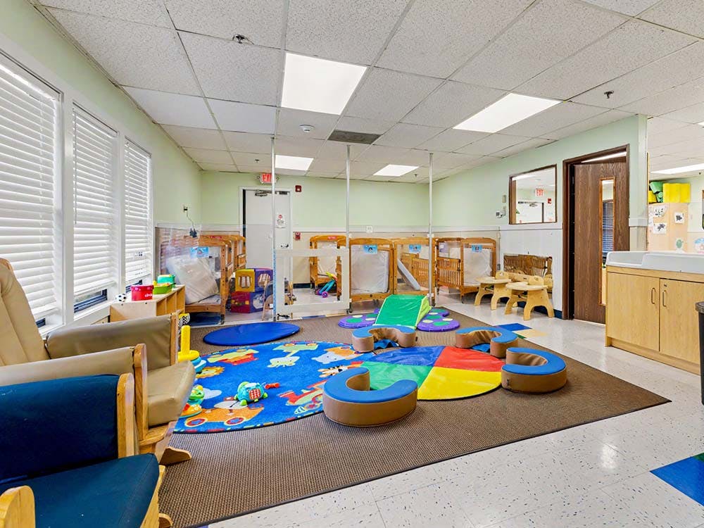 A colorful childcare classroom with cribs, play mats, and small tables