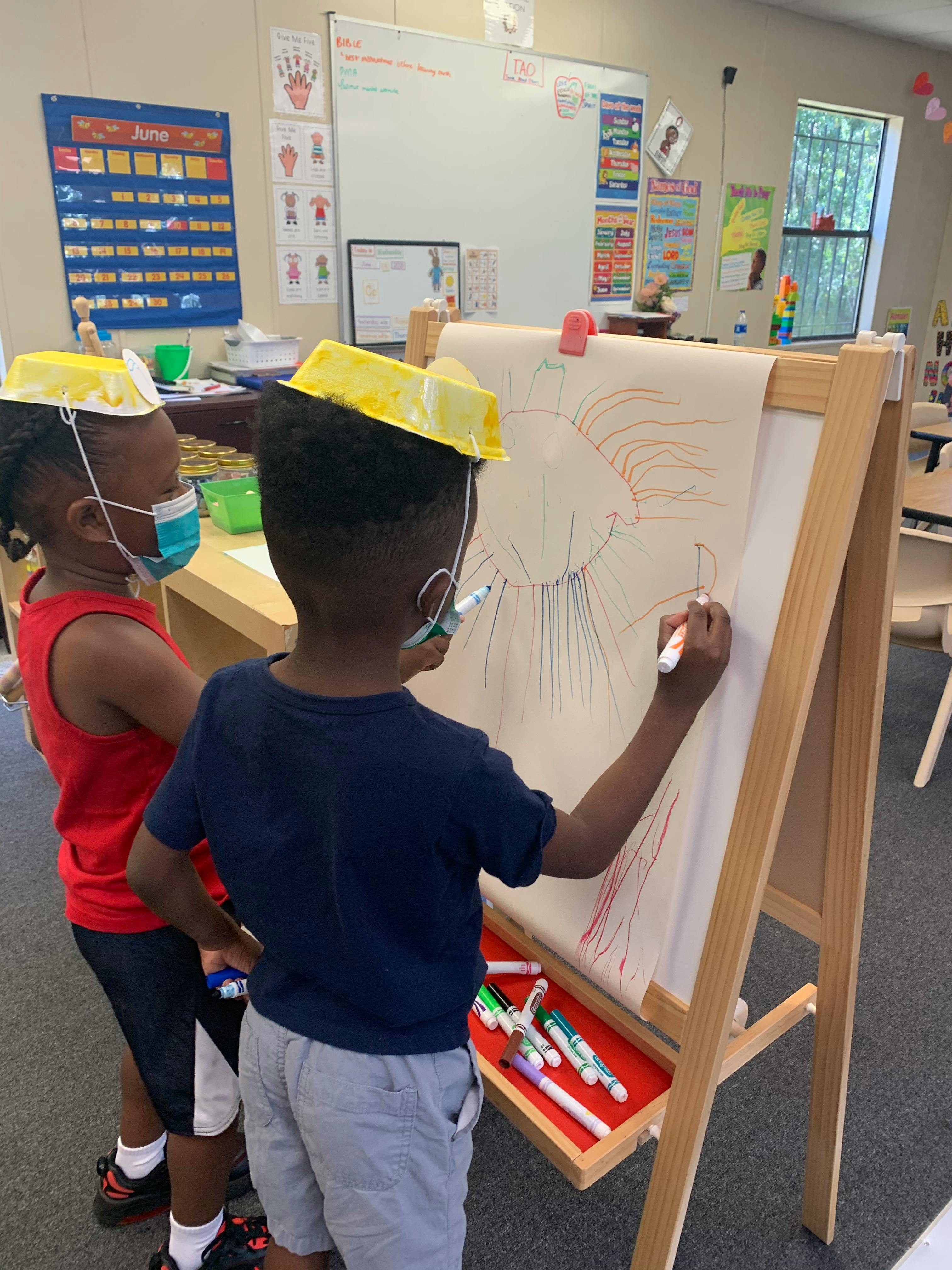 Two children in masks drawing on an easel in a classroom