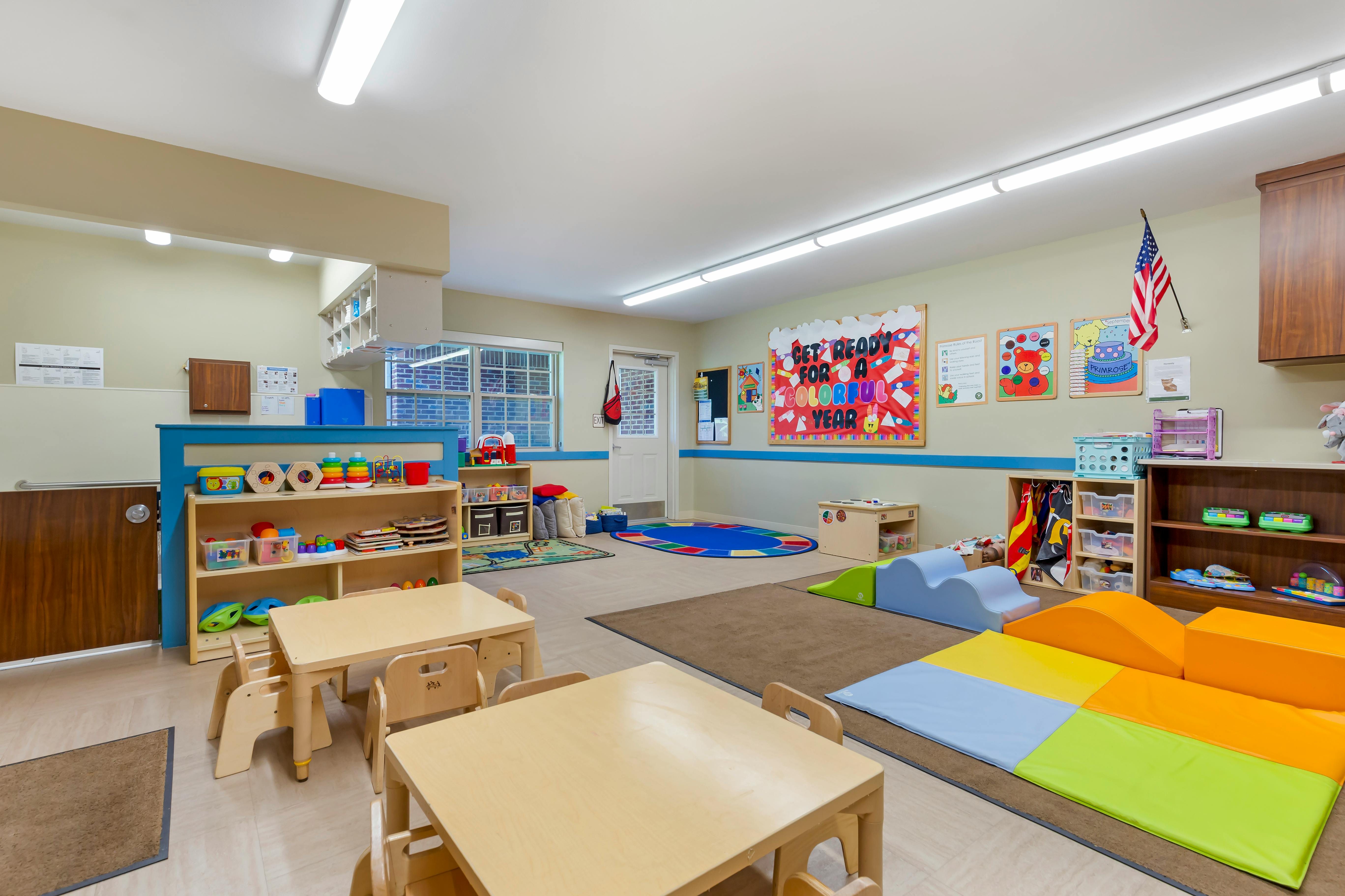 A colorful classroom with small tables and chairs, shelves of toys, and soft play equipment