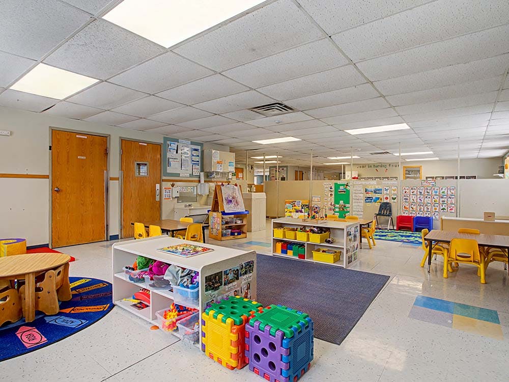 Empty classroom with small tables and chairs, shelves with toys, and posters