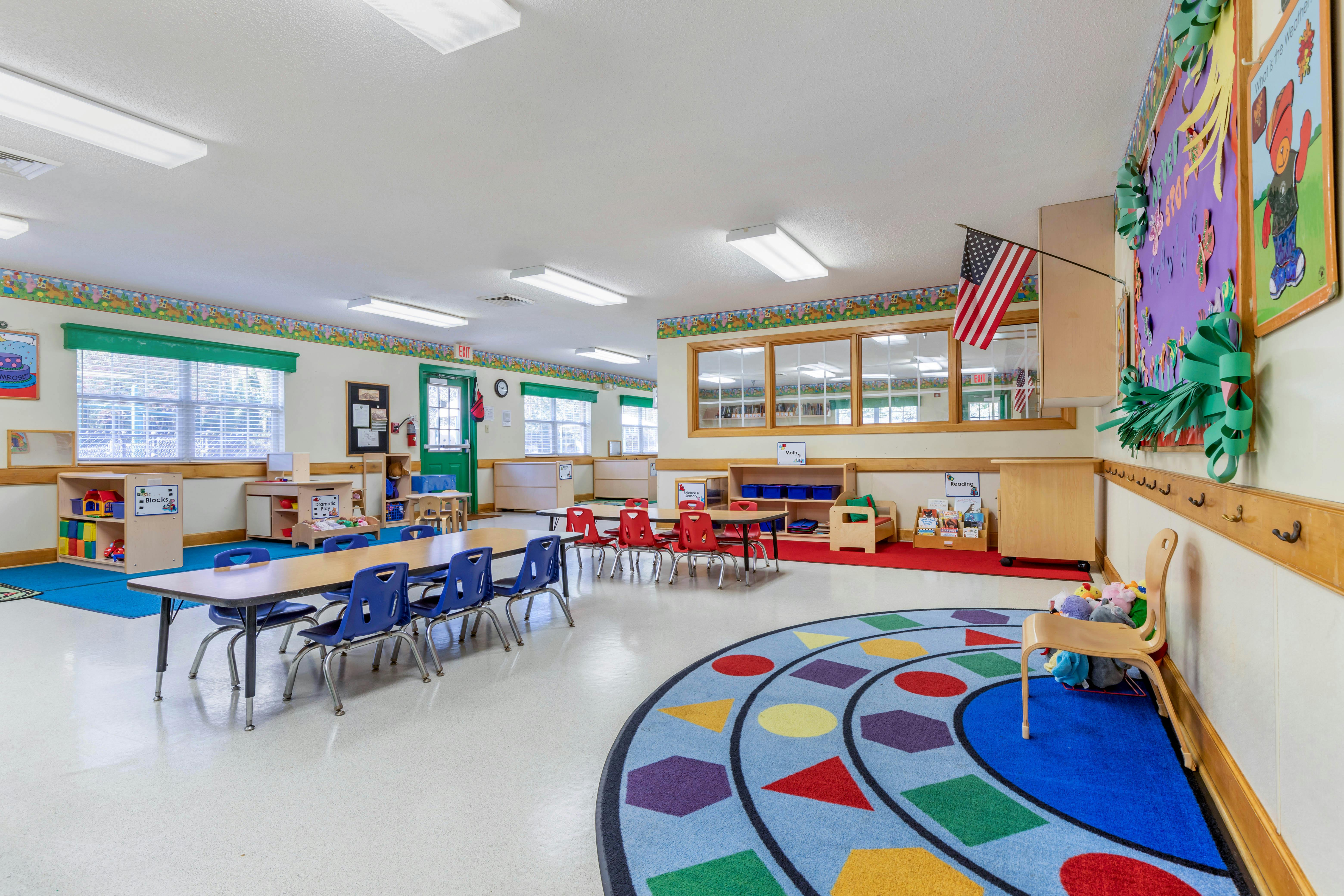 Empty classroom with small tables and chairs, colorful rugs, and play areas