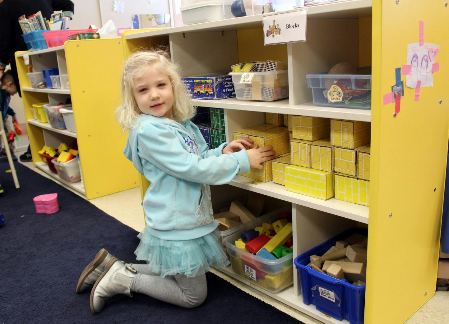 Child kneeling and reaching for blocks on a shelf in a classroom