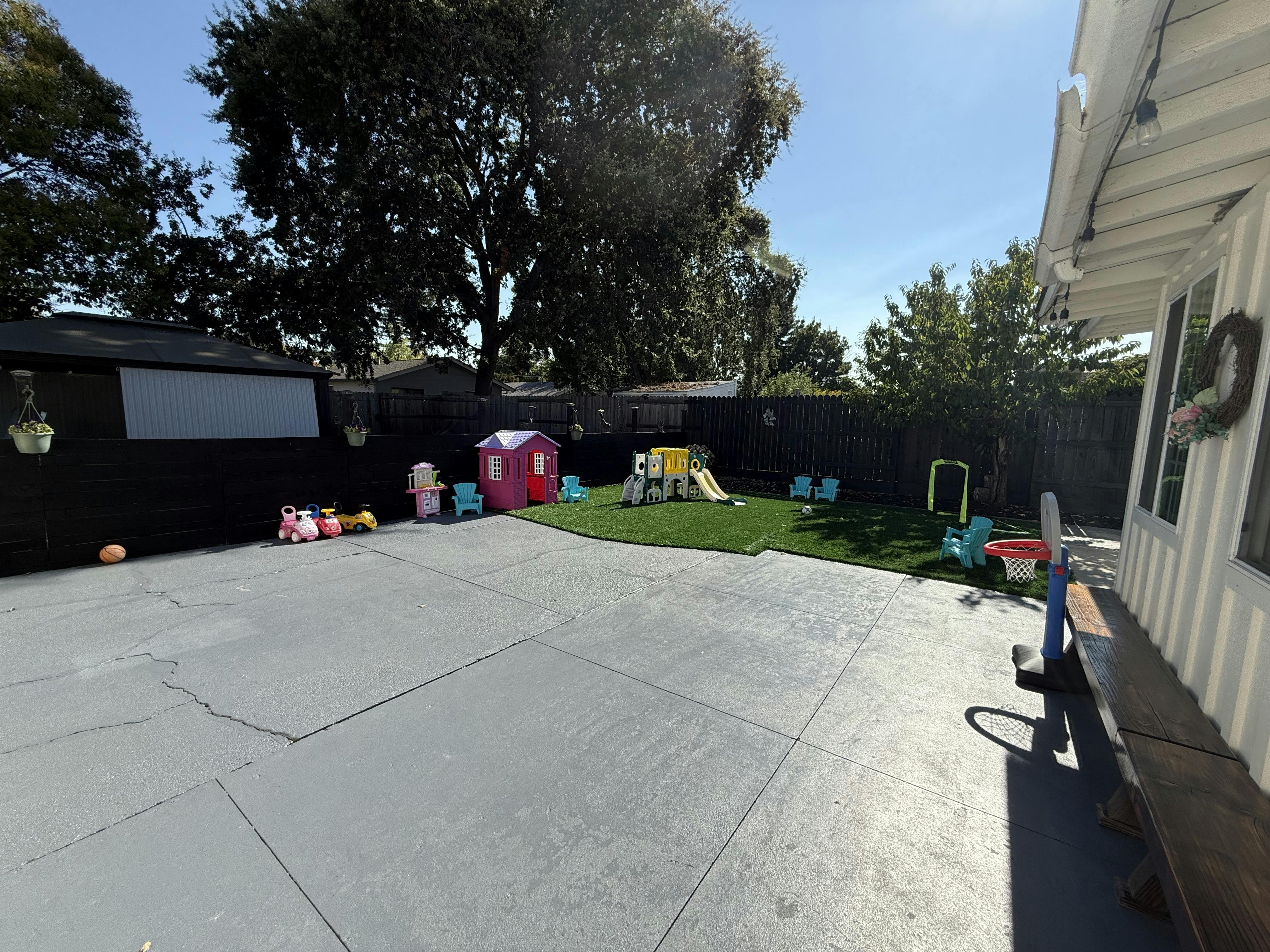 Outdoor play area with a small slide, playhouse, and toys on grass and pavement