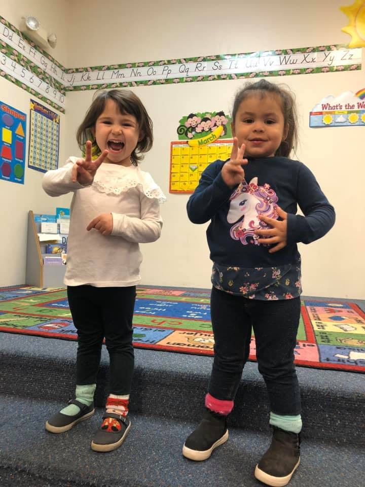 Two children standing in a classroom making peace signs with their hands