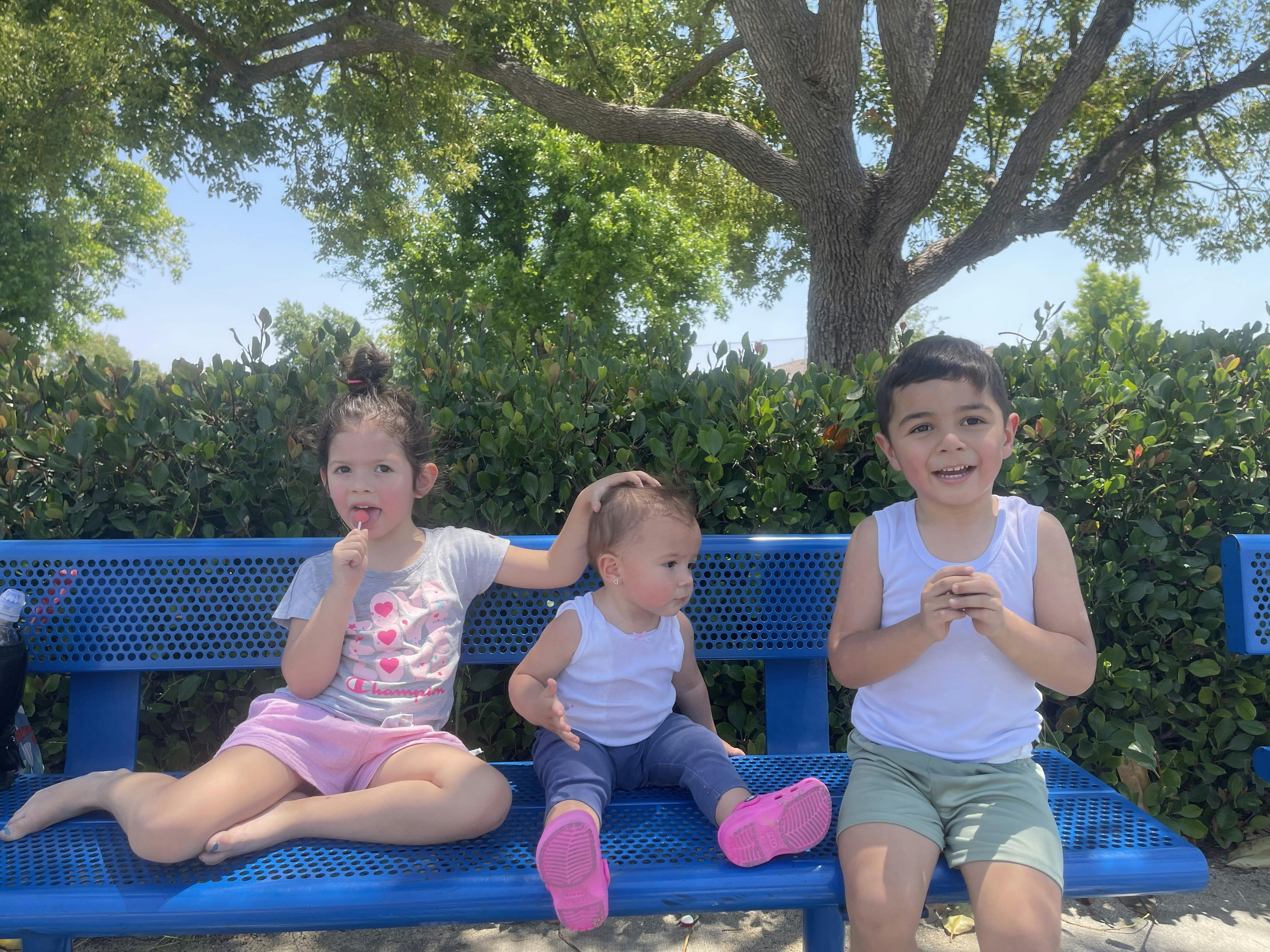 Three children sitting on a blue metal bench outdoors