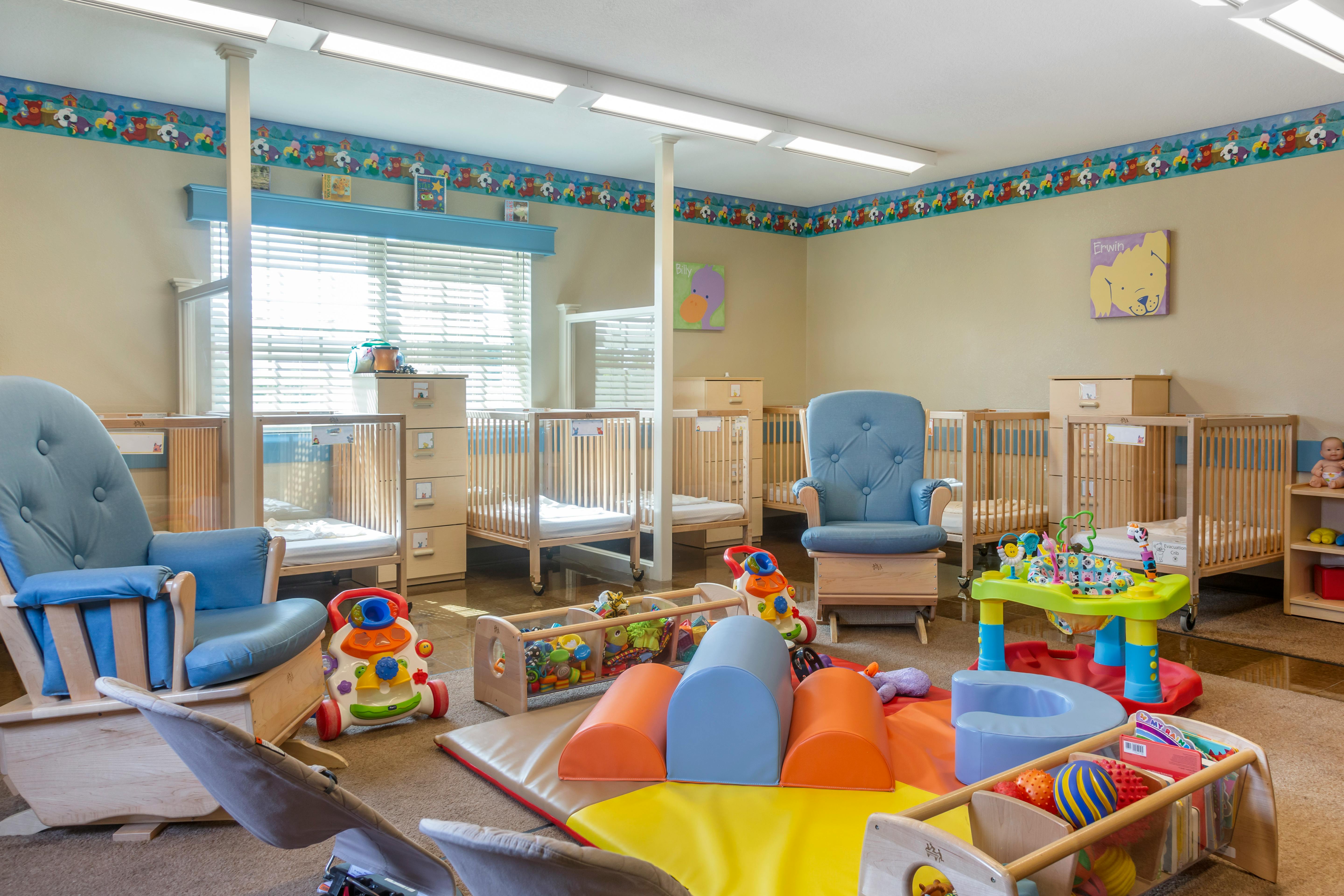 A childcare room with several cribs, two blue rocking chairs, and infant play equipment