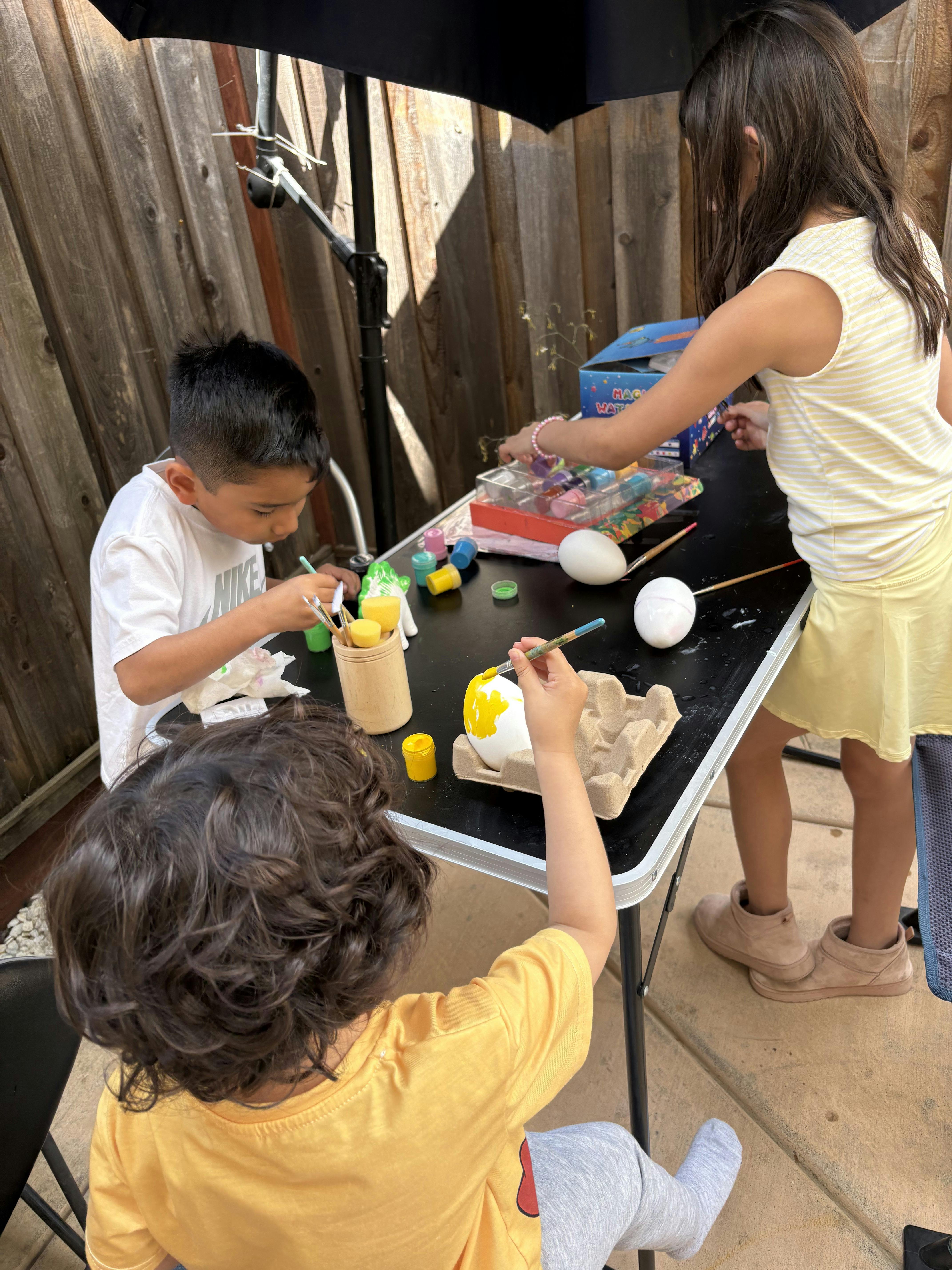 Three children painting Easter eggs on a black table outdoors