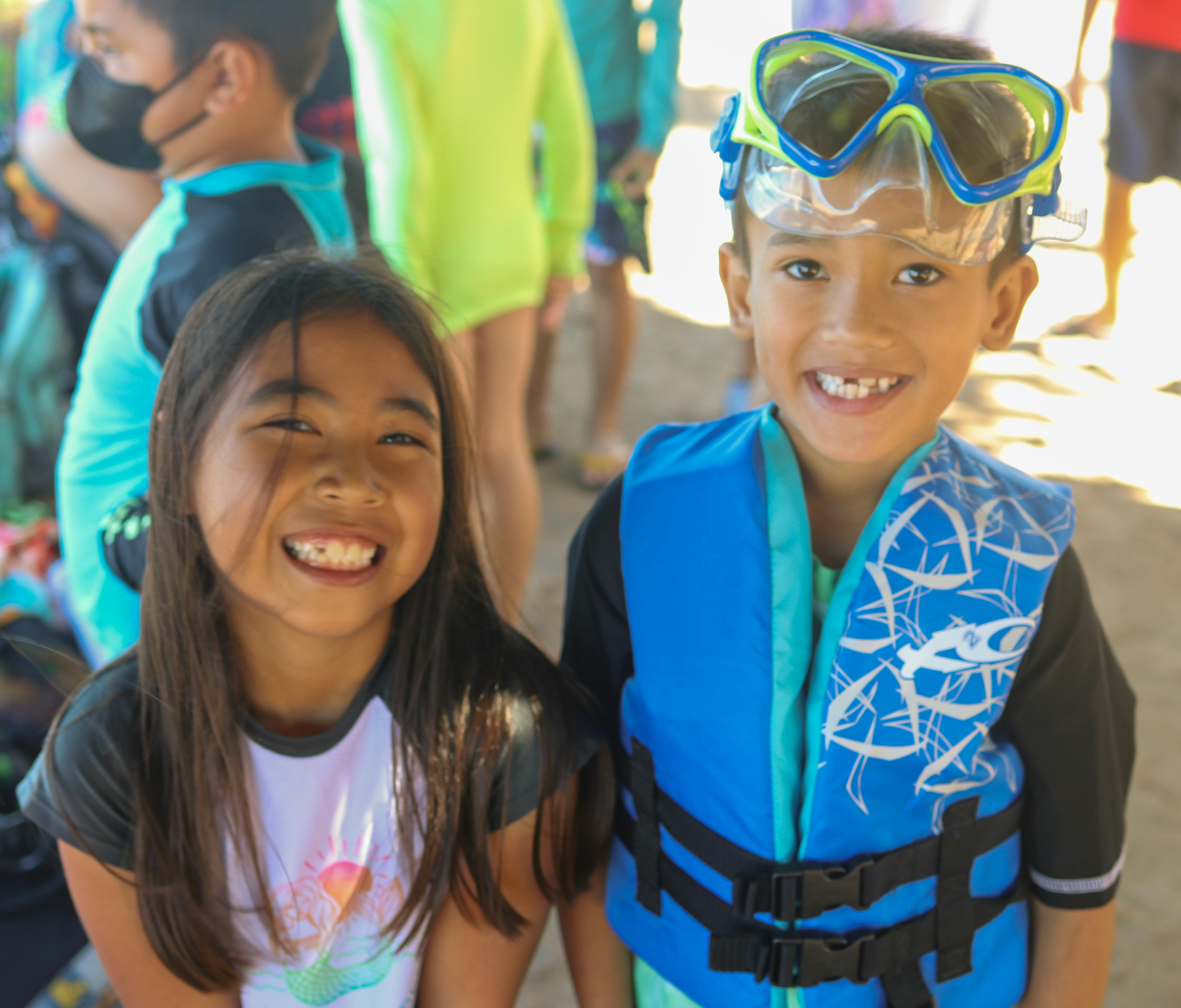 Two children smiling, one wearing a blue life vest and goggles