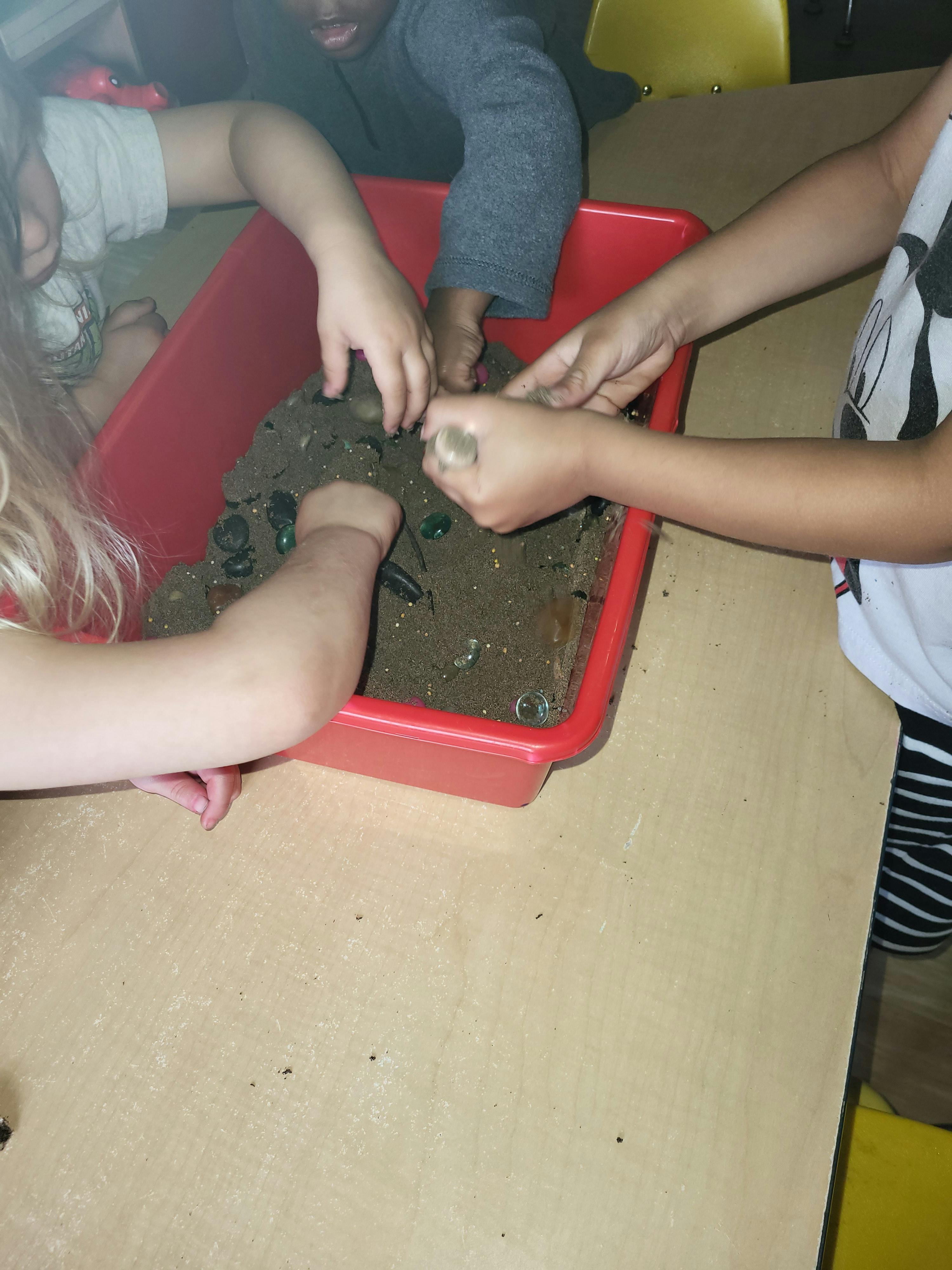 Children playing with sand and small objects in a red bin