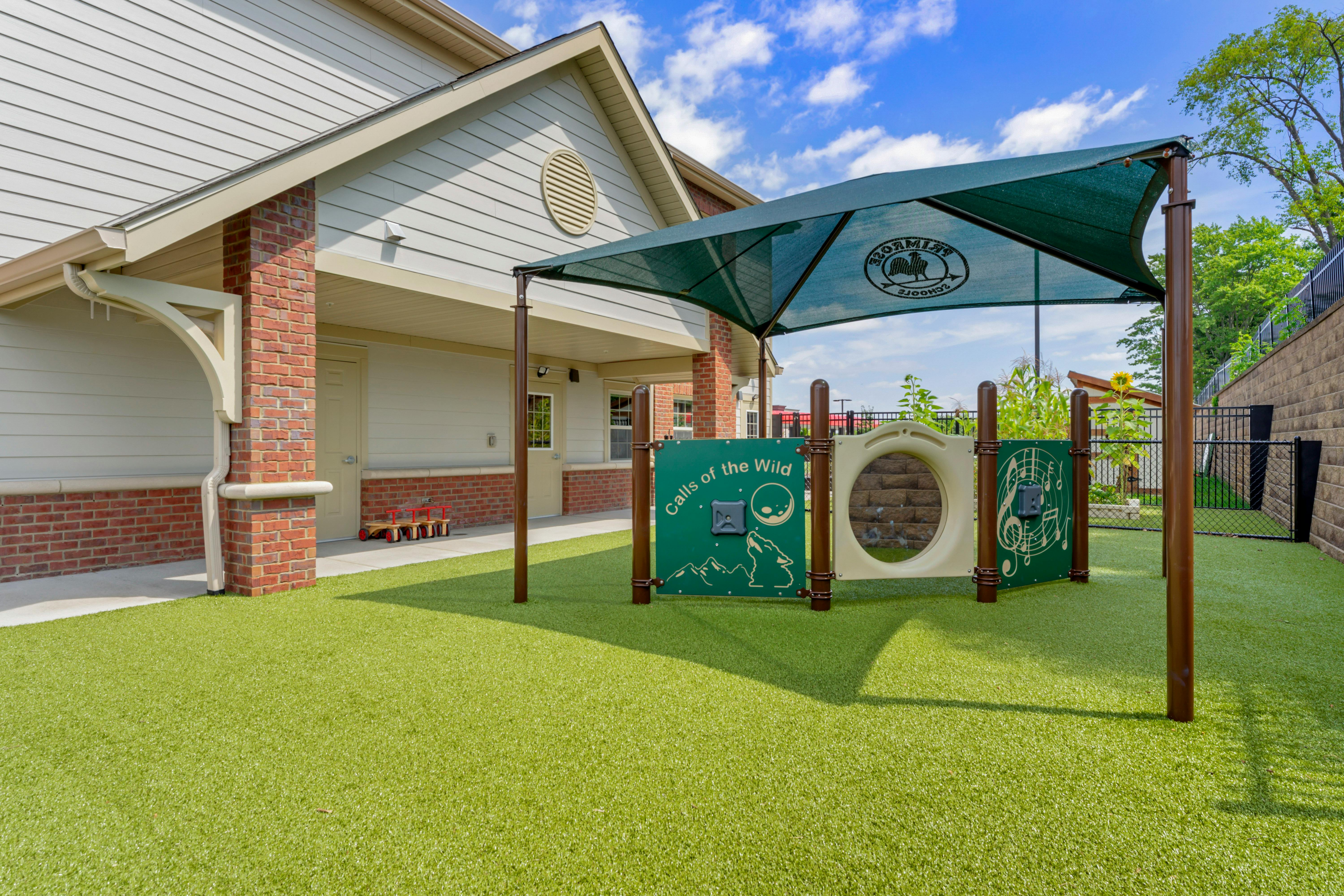 Outdoor playground with green artificial turf, a shaded play structure, and a building