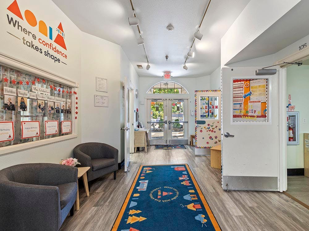 Hallway with a blue rug, two gray chairs, a bulletin board, and glass doors