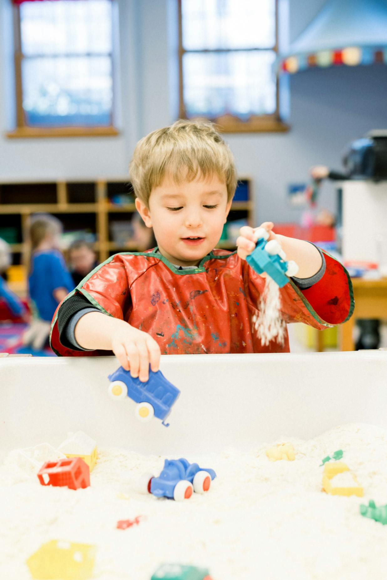Child in a red smock playing with toy cars in a sand table