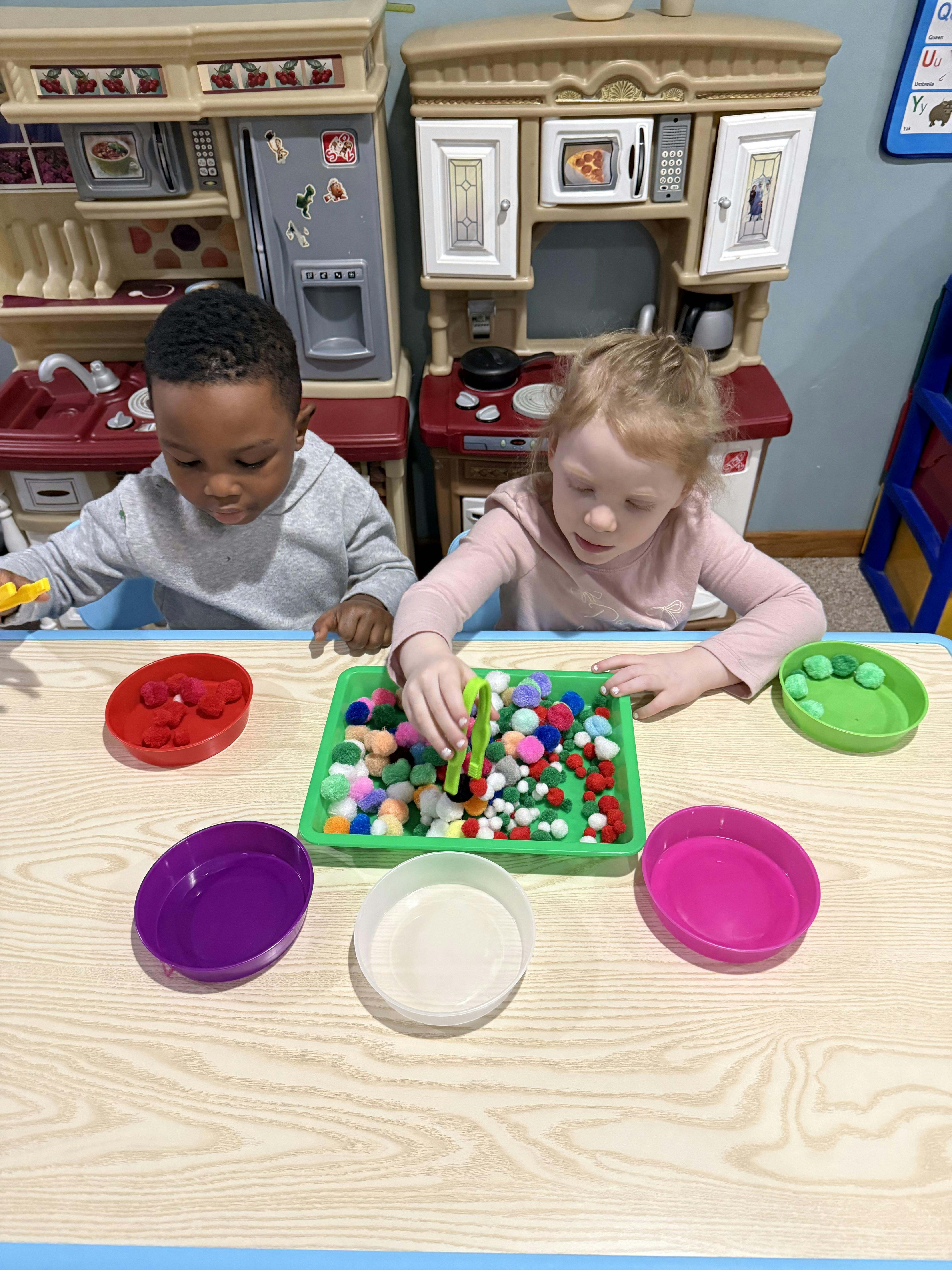 Two children playing with colorful pom-poms and tongs at a table