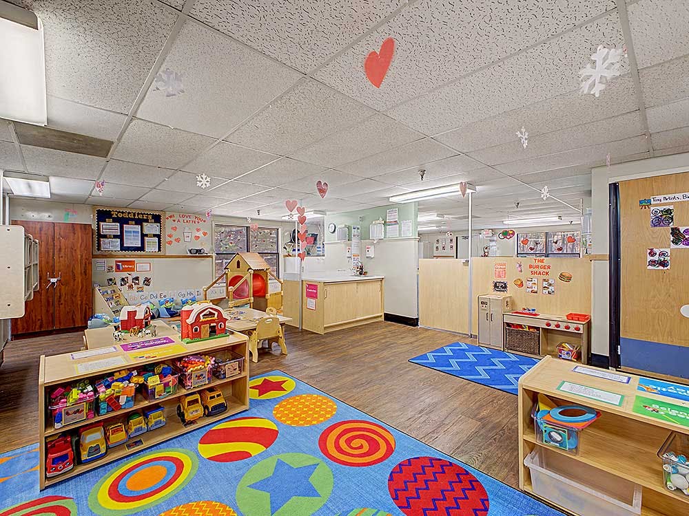 Brightly lit children's classroom with colorful rug, play kitchen, and toy shelf