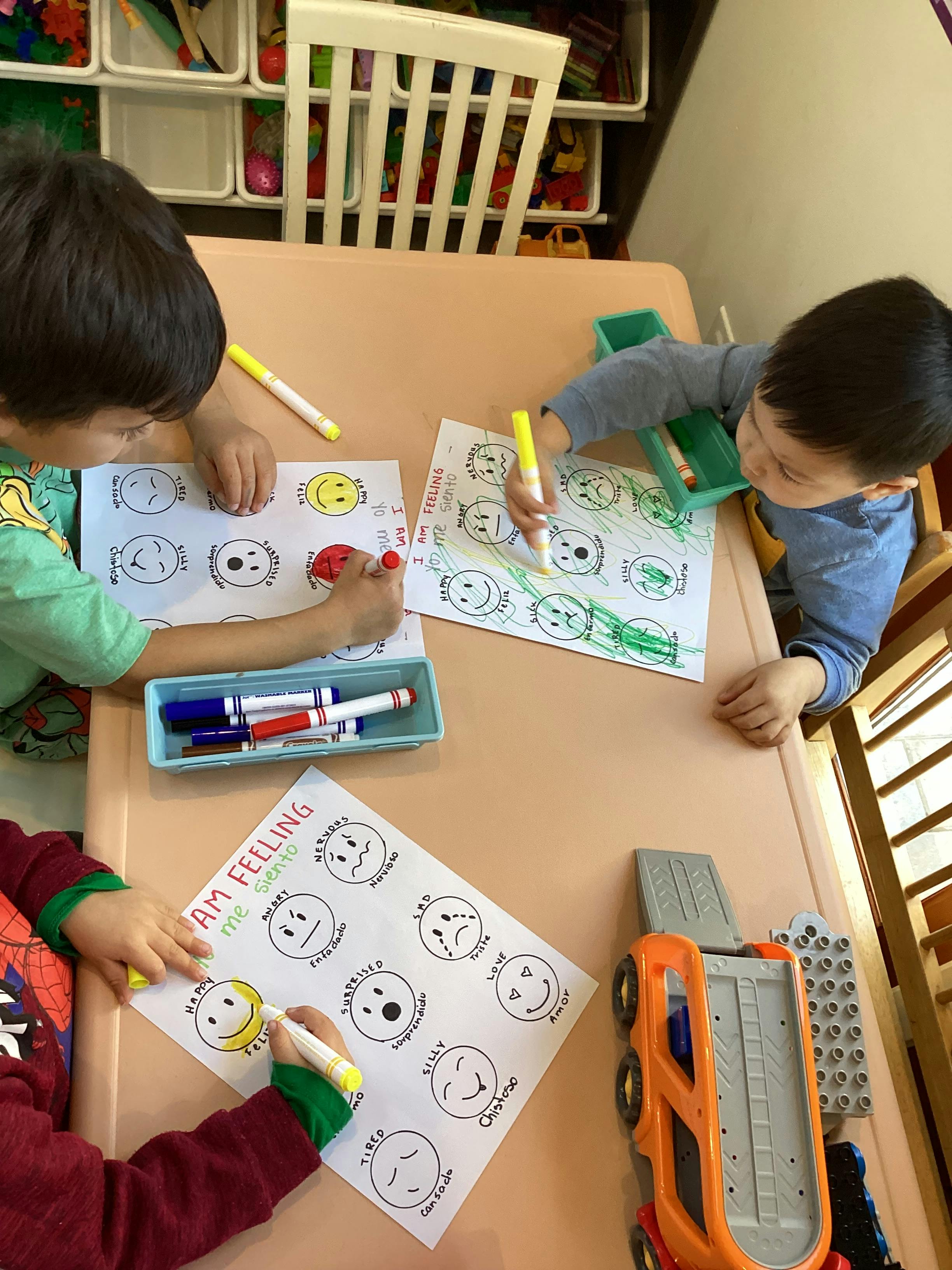 Three children coloring emoji faces on paper at a table
