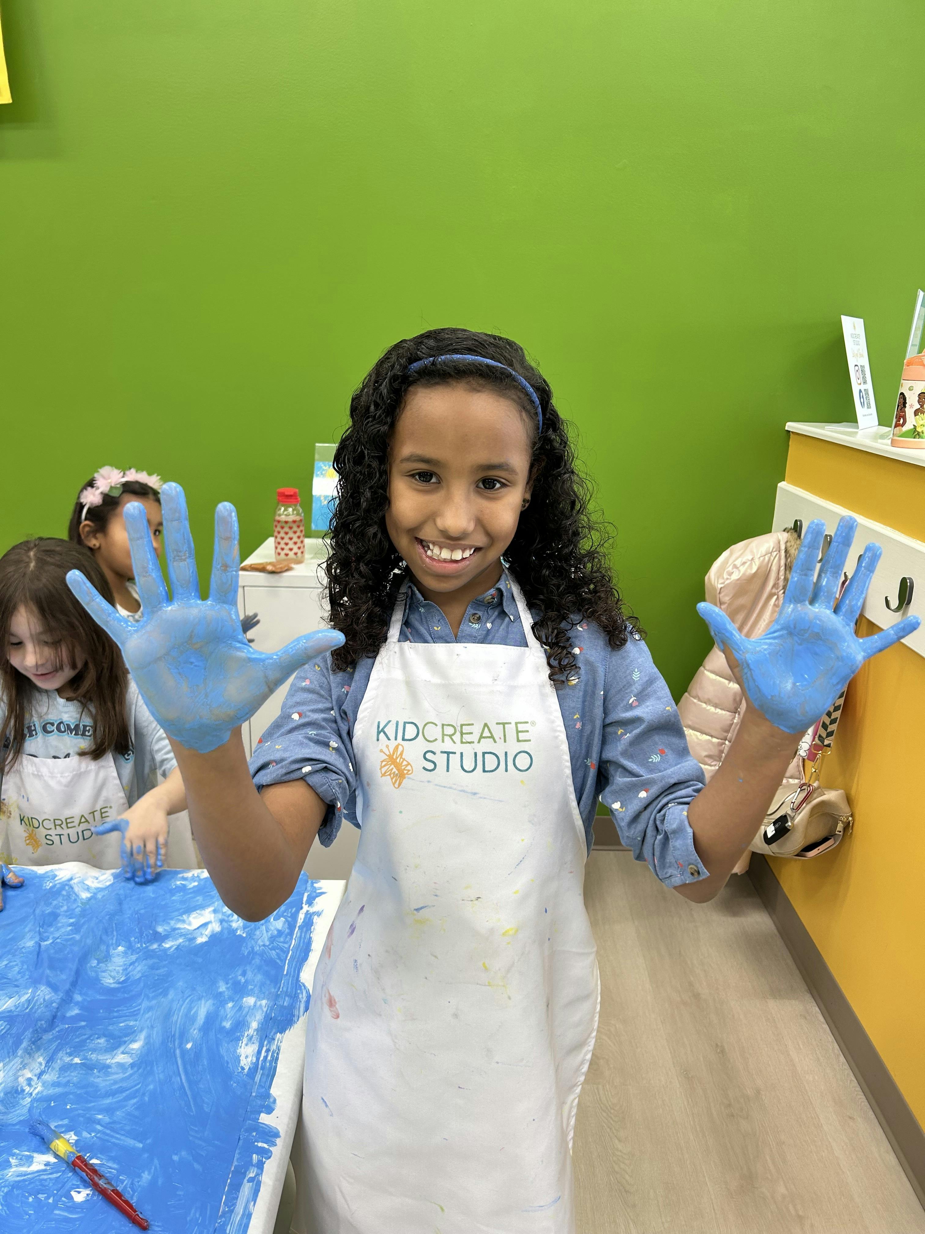 Child in an apron showing hands covered in blue paint, with another child in the background