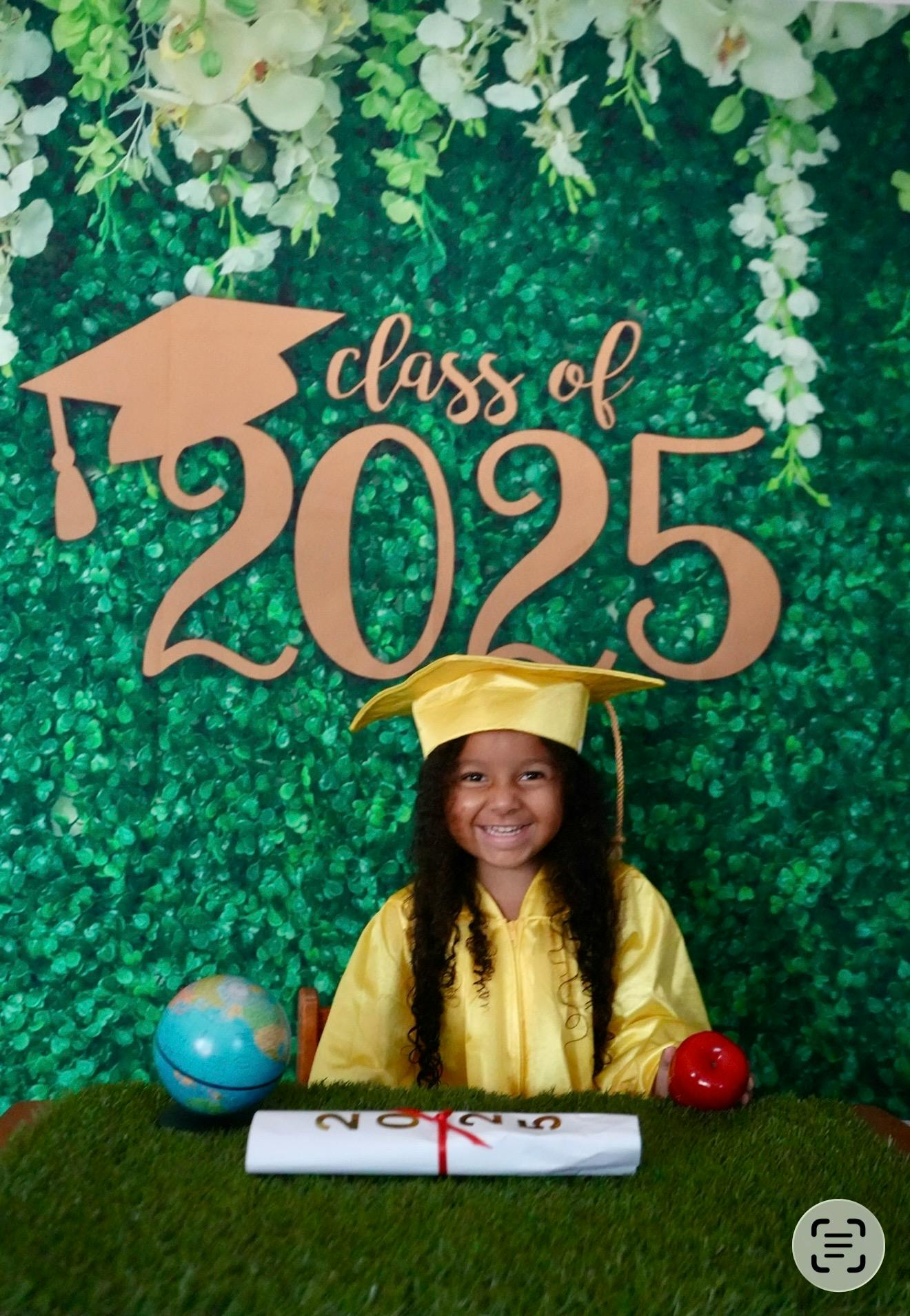 Child wearing graduation cap and gown in front of a class of 2025 sign