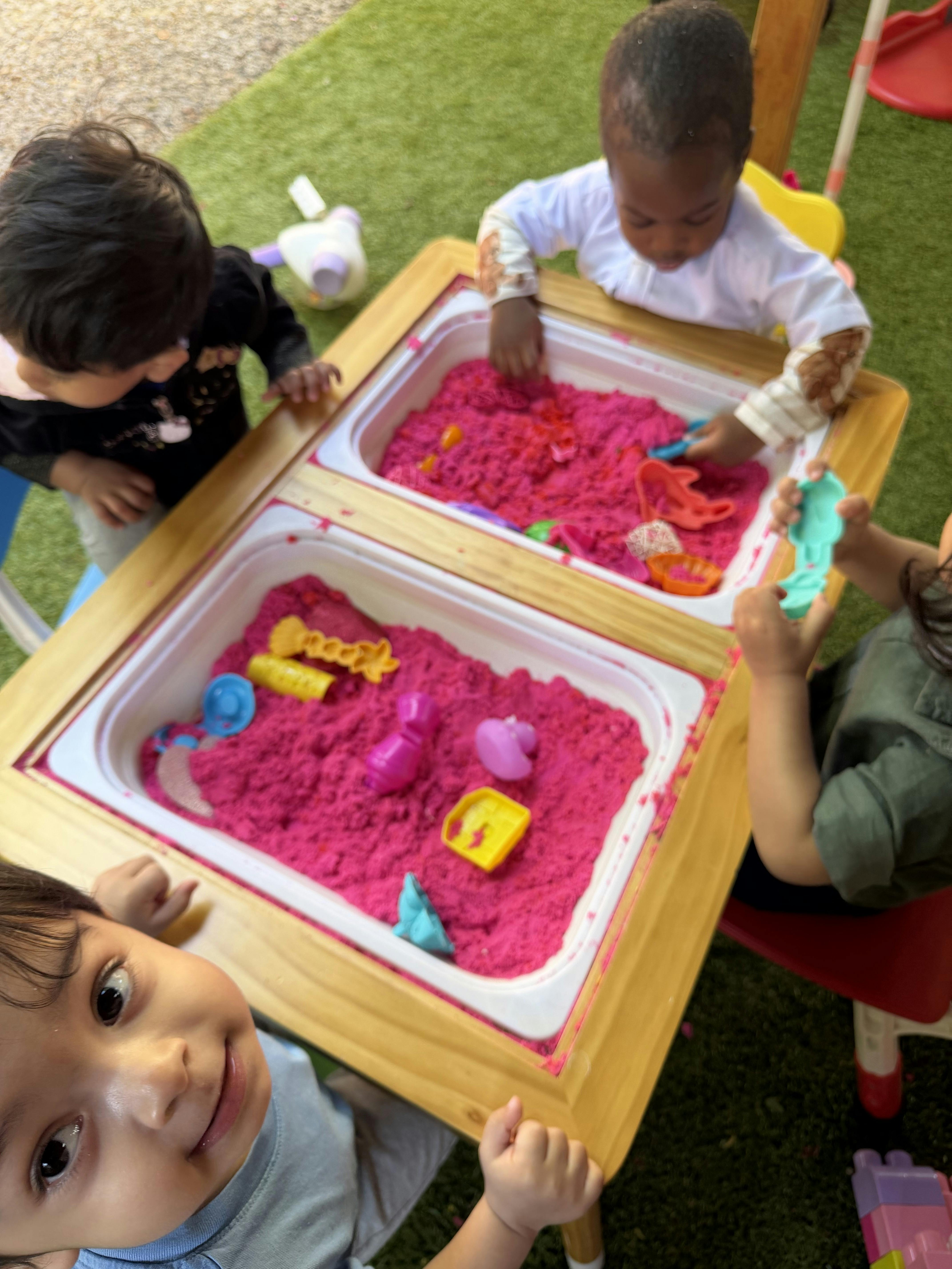 Four children playing with pink kinetic sand and toys at a table