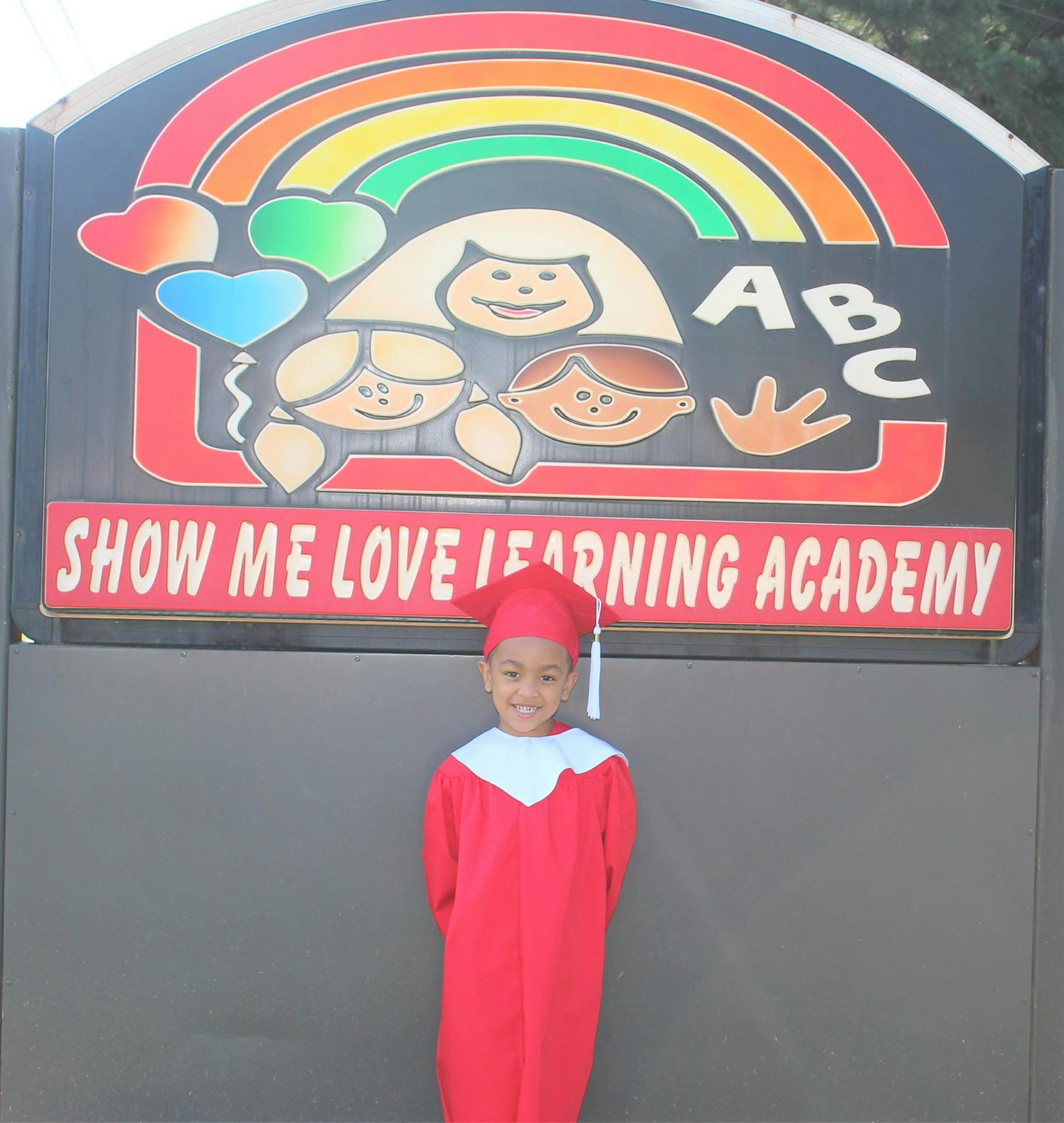 Child in red graduation cap and gown in front of a sign for a learning academy sign