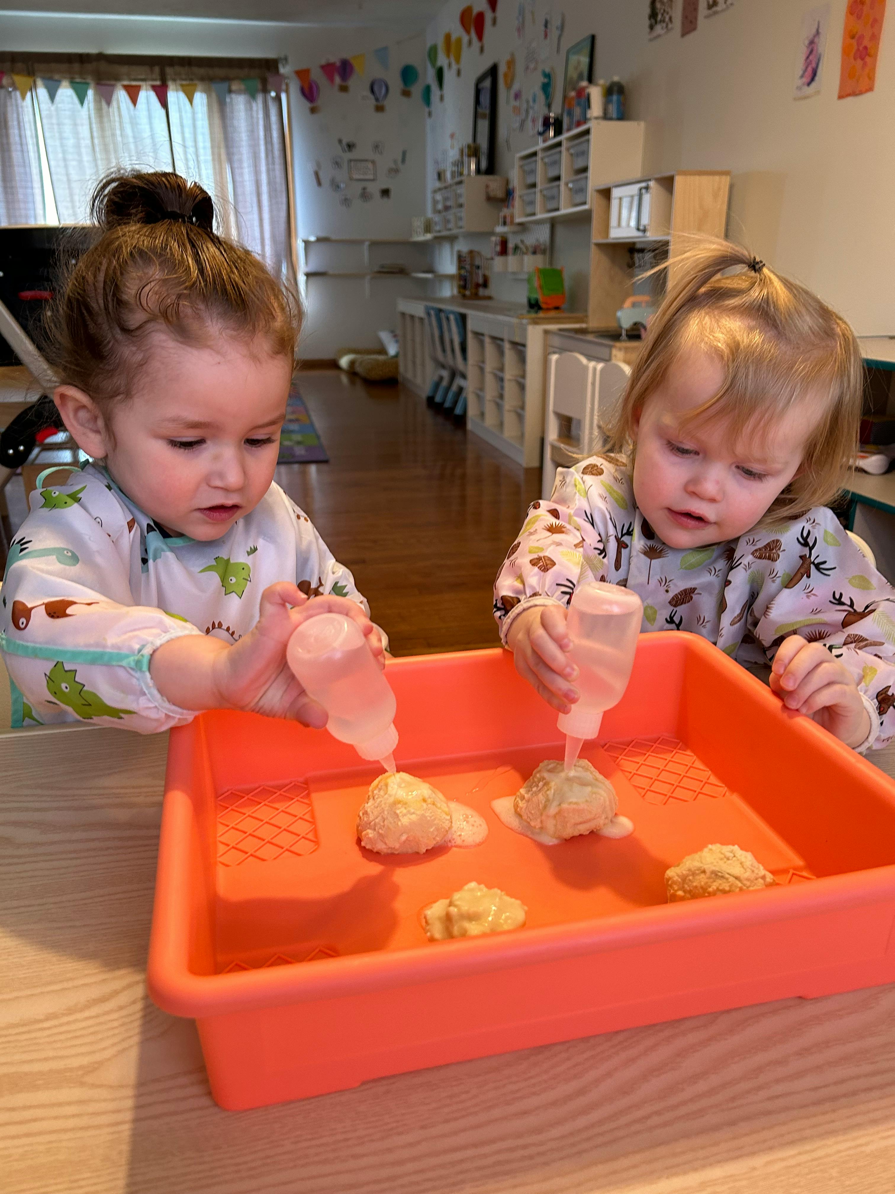 Two children squeezing liquid from bottles onto objects in a tray