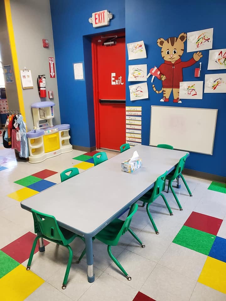 Classroom with a long table and green chairs, colorful floor tiles, and a red exit door