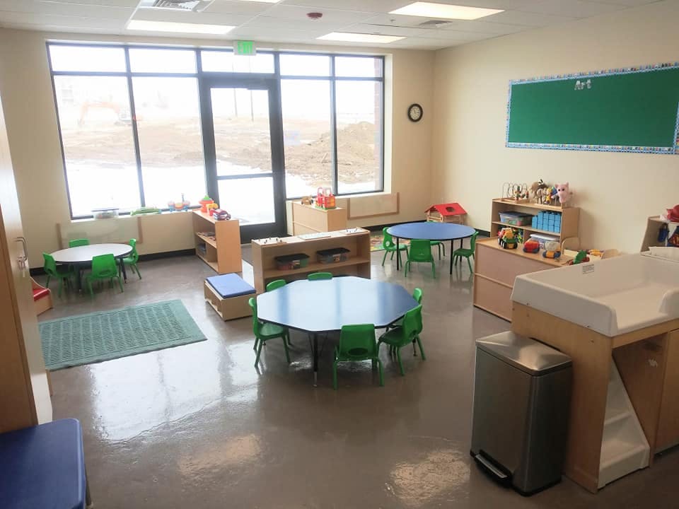 Empty classroom with small tables and chairs, large windows, and a green chalkboard
