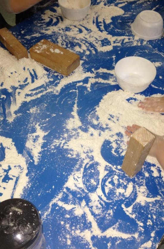 Hands playing with flour on a blue surface with wooden blocks and bowls