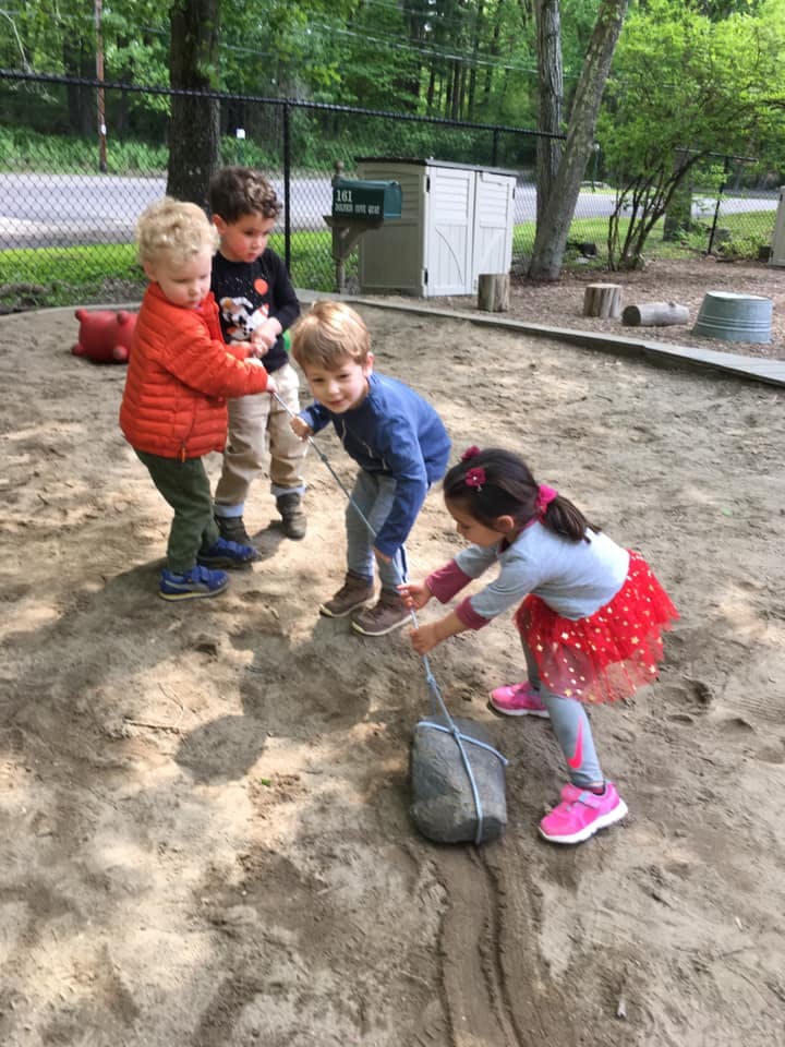 Four children playing in a sandbox, one pulling a rock with a rope