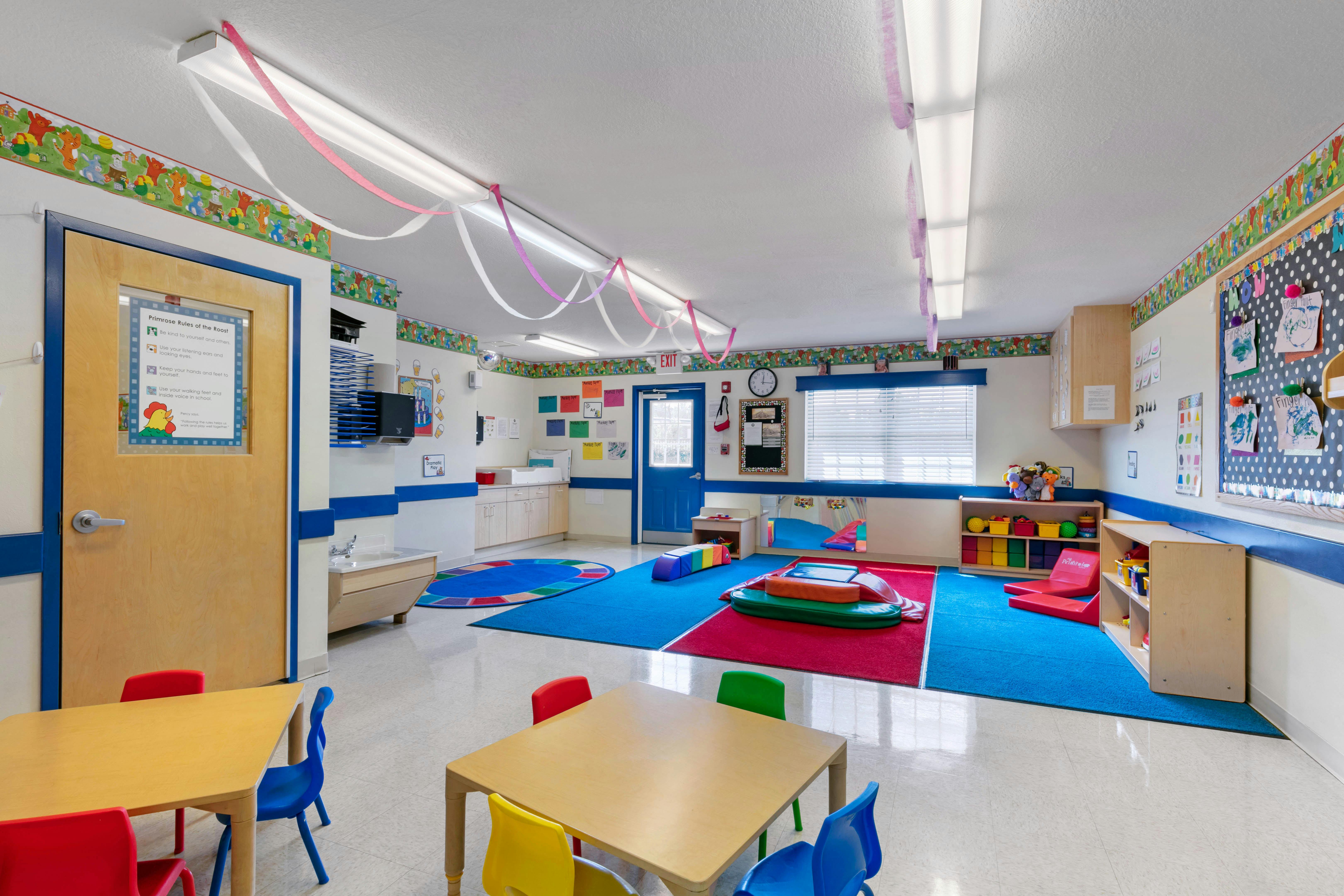 A colorful empty classroom with small tables and chairs, bright rugs, and an exit door