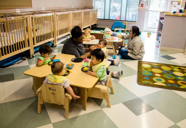 Adults and children sitting at tables in a room with cribs