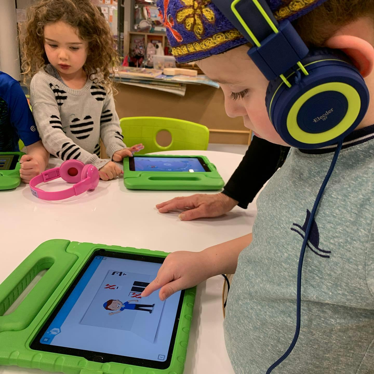 Child wearing headphones using a tablet, another child watching