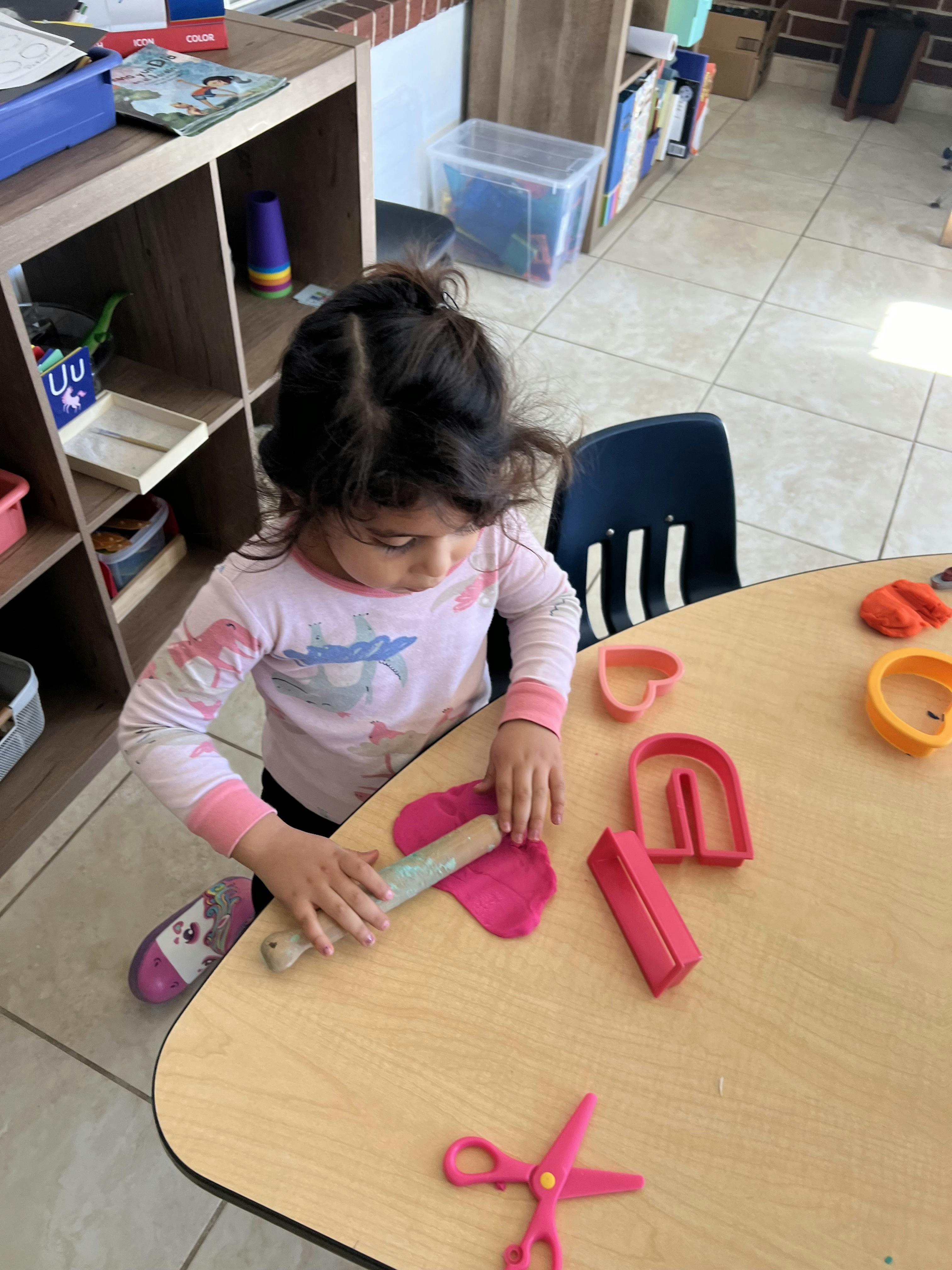 Child rolling pink playdoh on a wooden table in a classroom