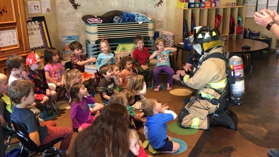 Person in firefighter gear kneeling in front of children in a classroom