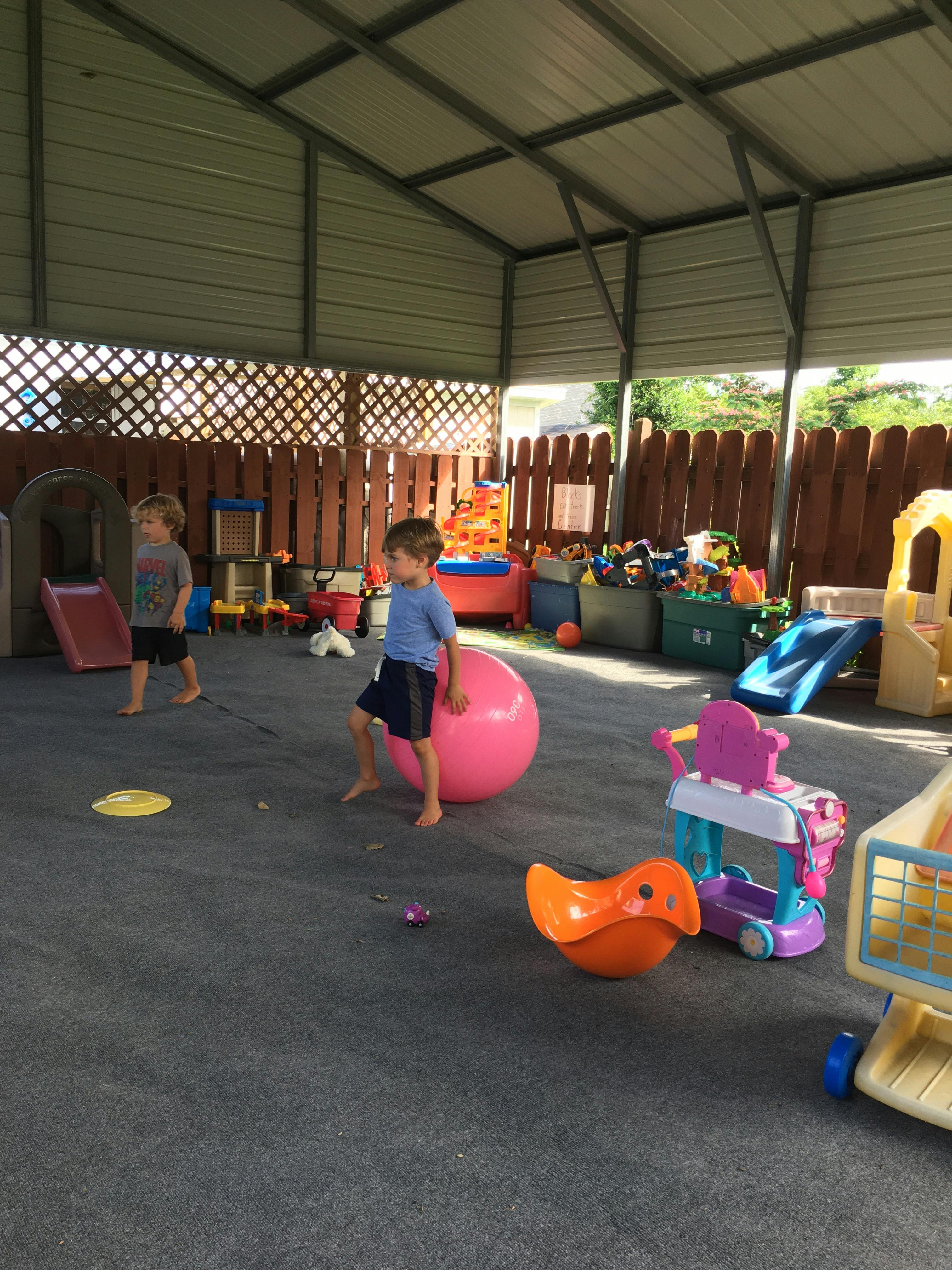 Two children playing with a large pink ball and other toys in a covered outdoor area