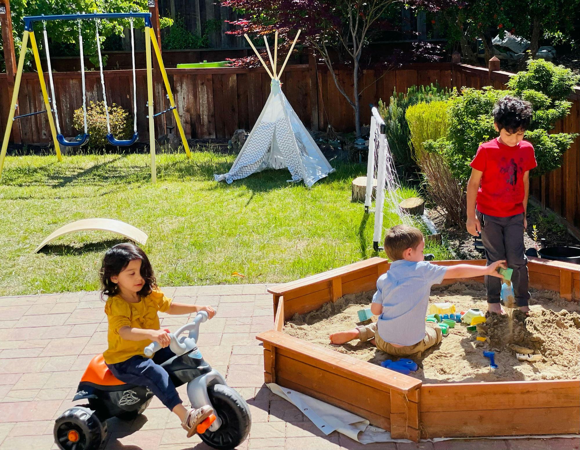 Three children playing in a backyard, one on a tricycle and two in a sandbox