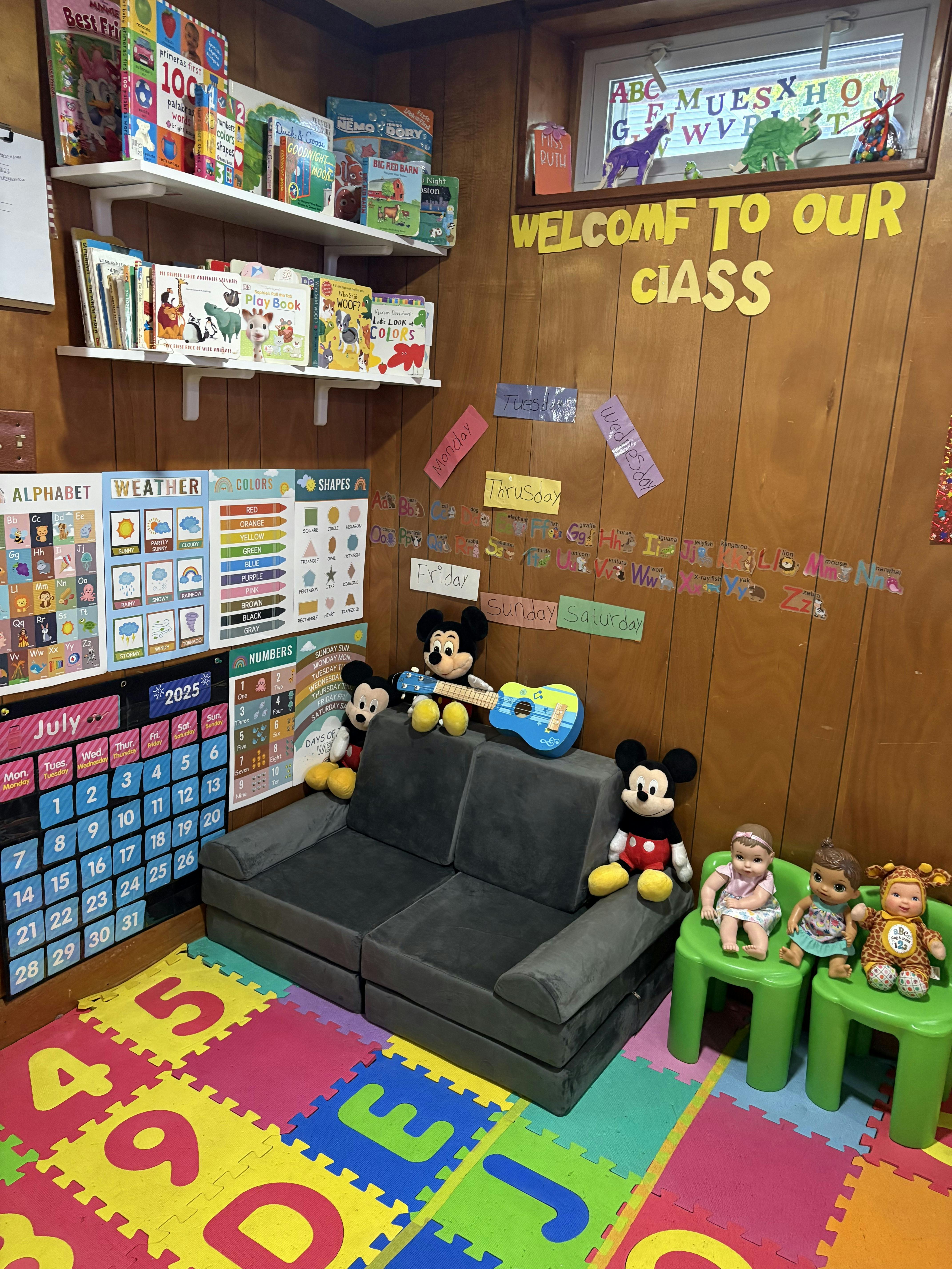 Classroom corner with bookshelves, educational charts, a small couch, and dolls