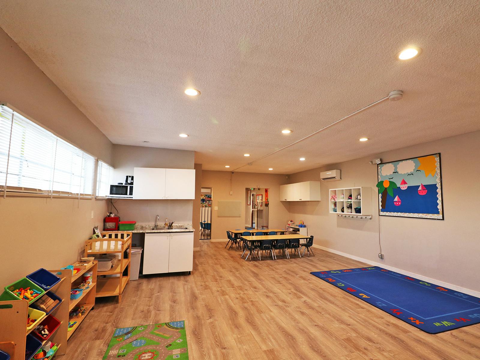 Empty classroom with small tables and chairs, a kitchen area, and a blue rug