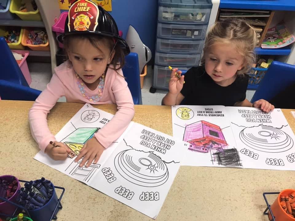 Two children coloring at a table, one wearing a firefighter's hat