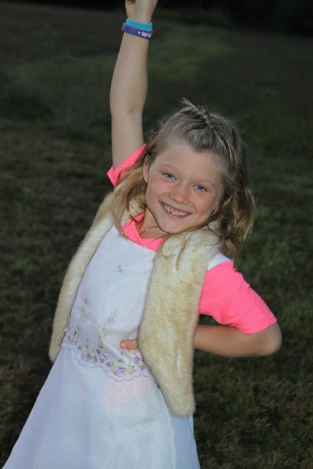 Child in a white dress and fur vest with arm raised