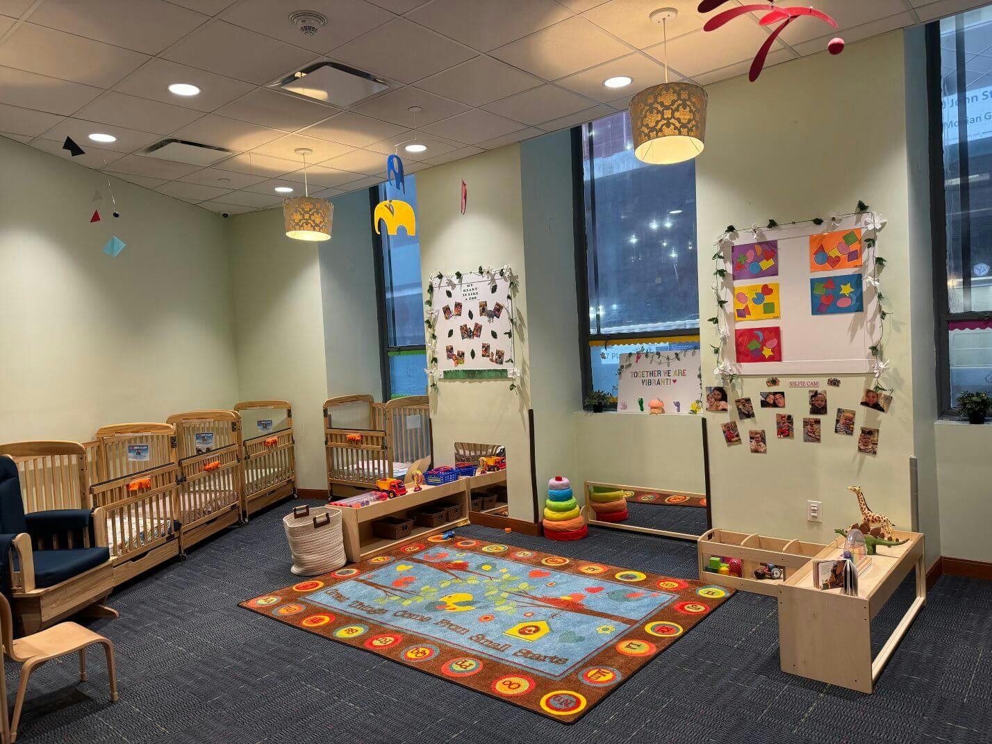 cribs in a brightly lit daycare room with a colorful rug and learning materials