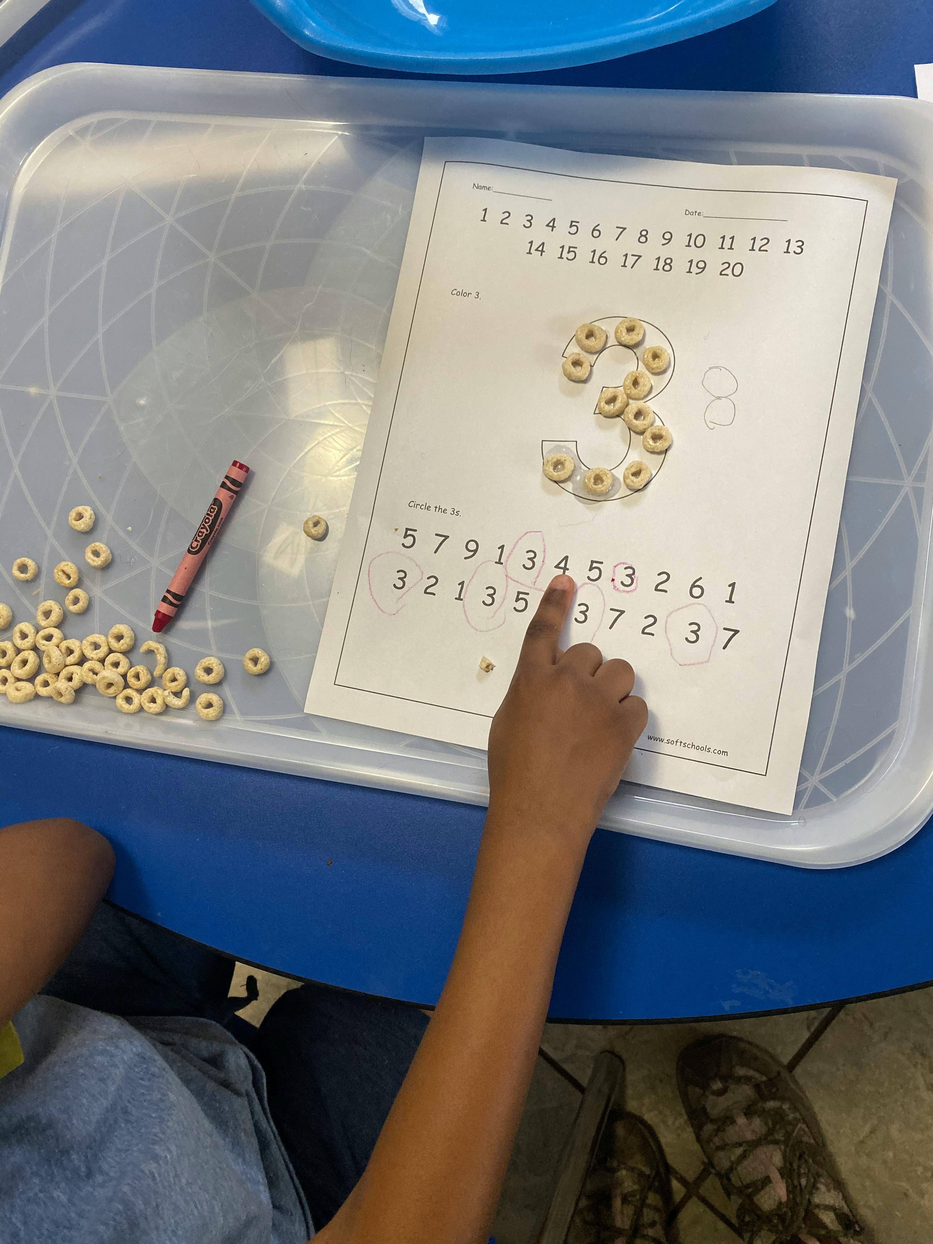 Child's hand pointing at a number worksheet with cereal pieces