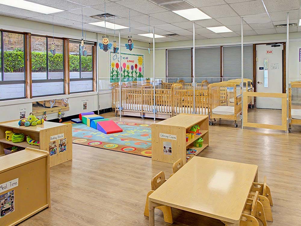 A brightly lit classroom with small tables, play mats, and several cribs