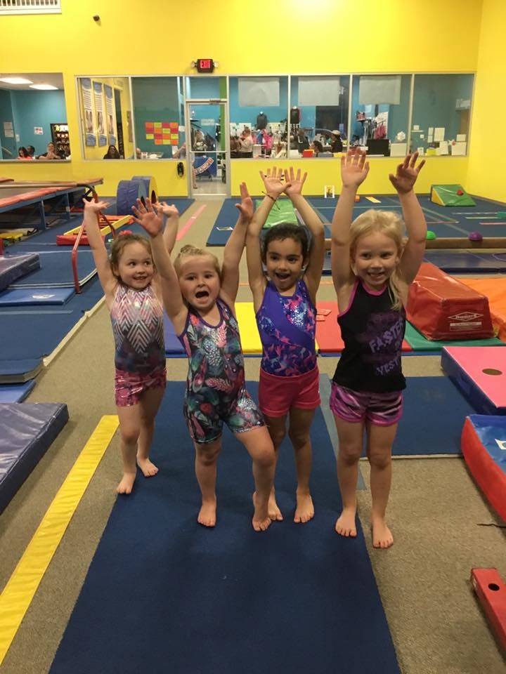 Four children in leotards with arms raised in a gymnastics gym