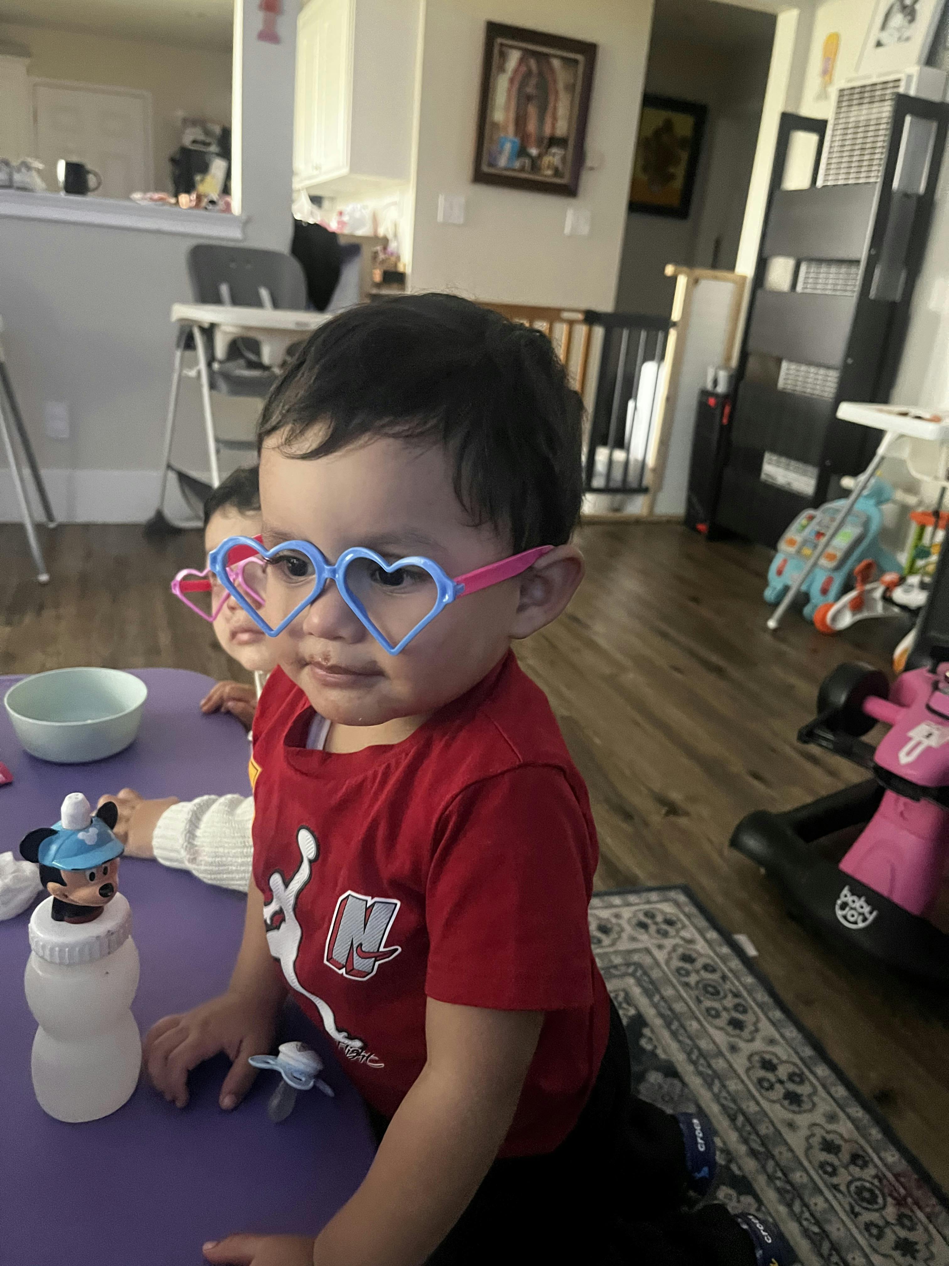 Child wearing blue heart-shaped glasses, another child in background