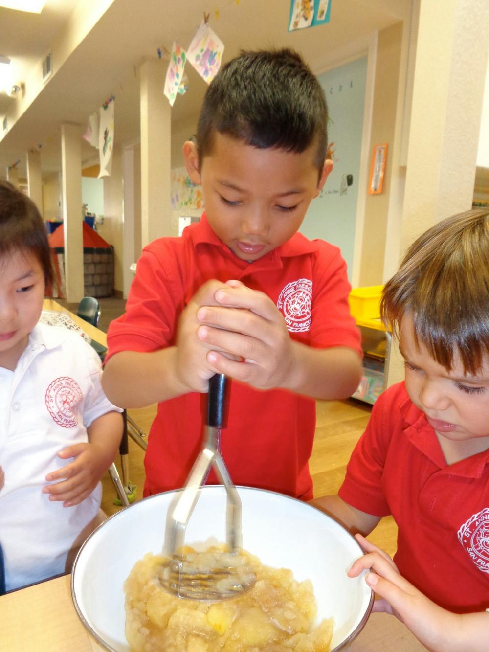 Child mashing fruit in a bowl while other children watch in a classroom