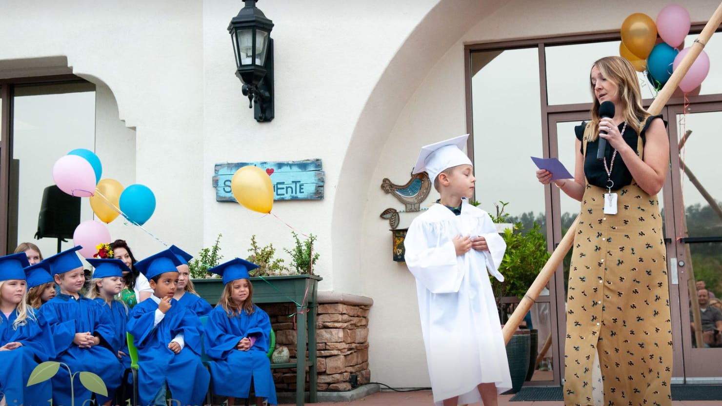 Adult speaking into microphone with children in graduation caps and white robes